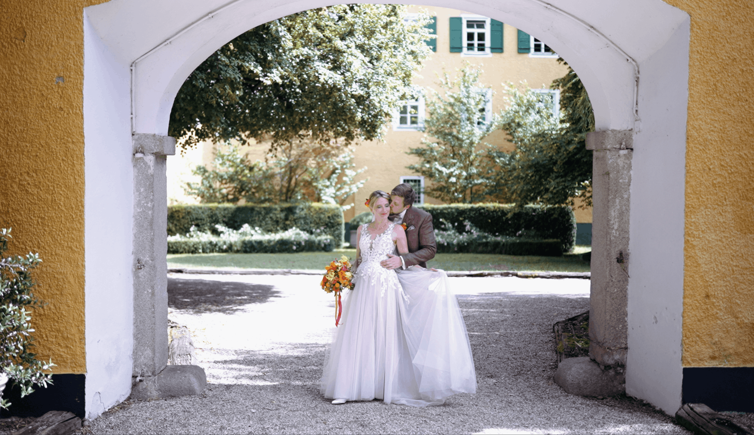 A bride in a white wedding dress and a groom in a suit are standing together under an archway, with a yellow building and green trees in the background.