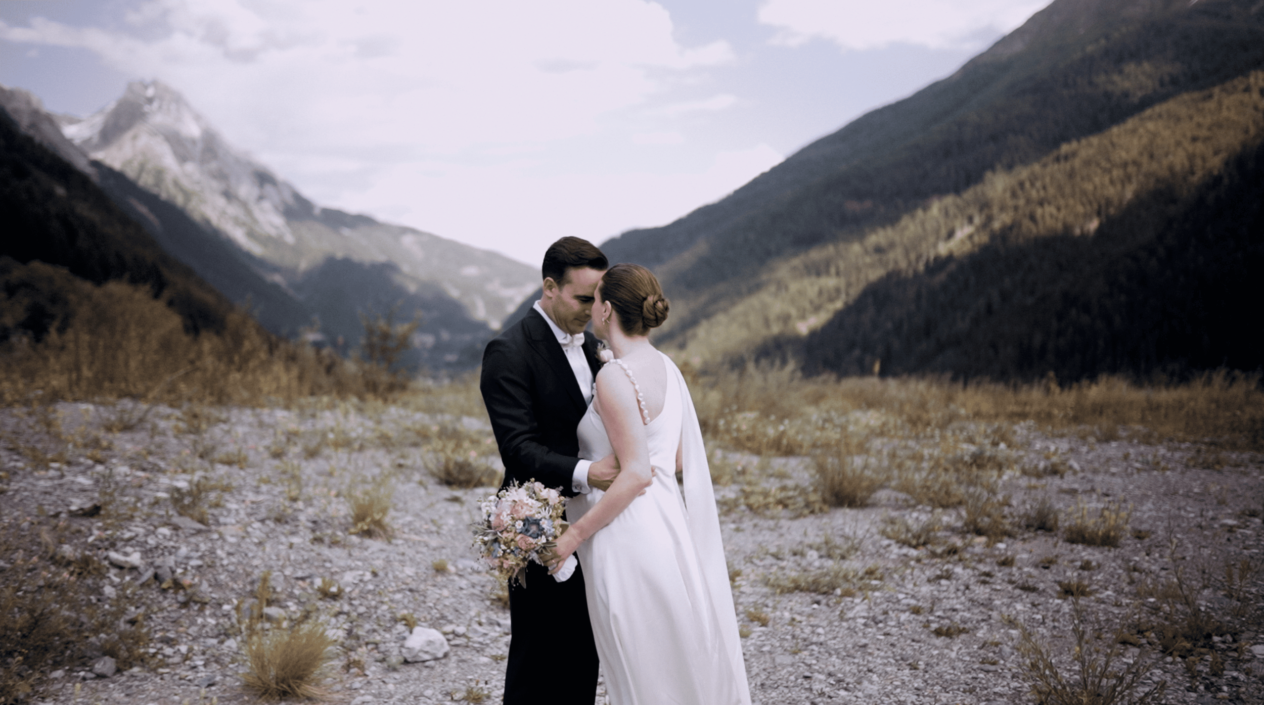 A bride and groom share a kiss in a mountain valley with rugged hills and a cloudy sky in the background.