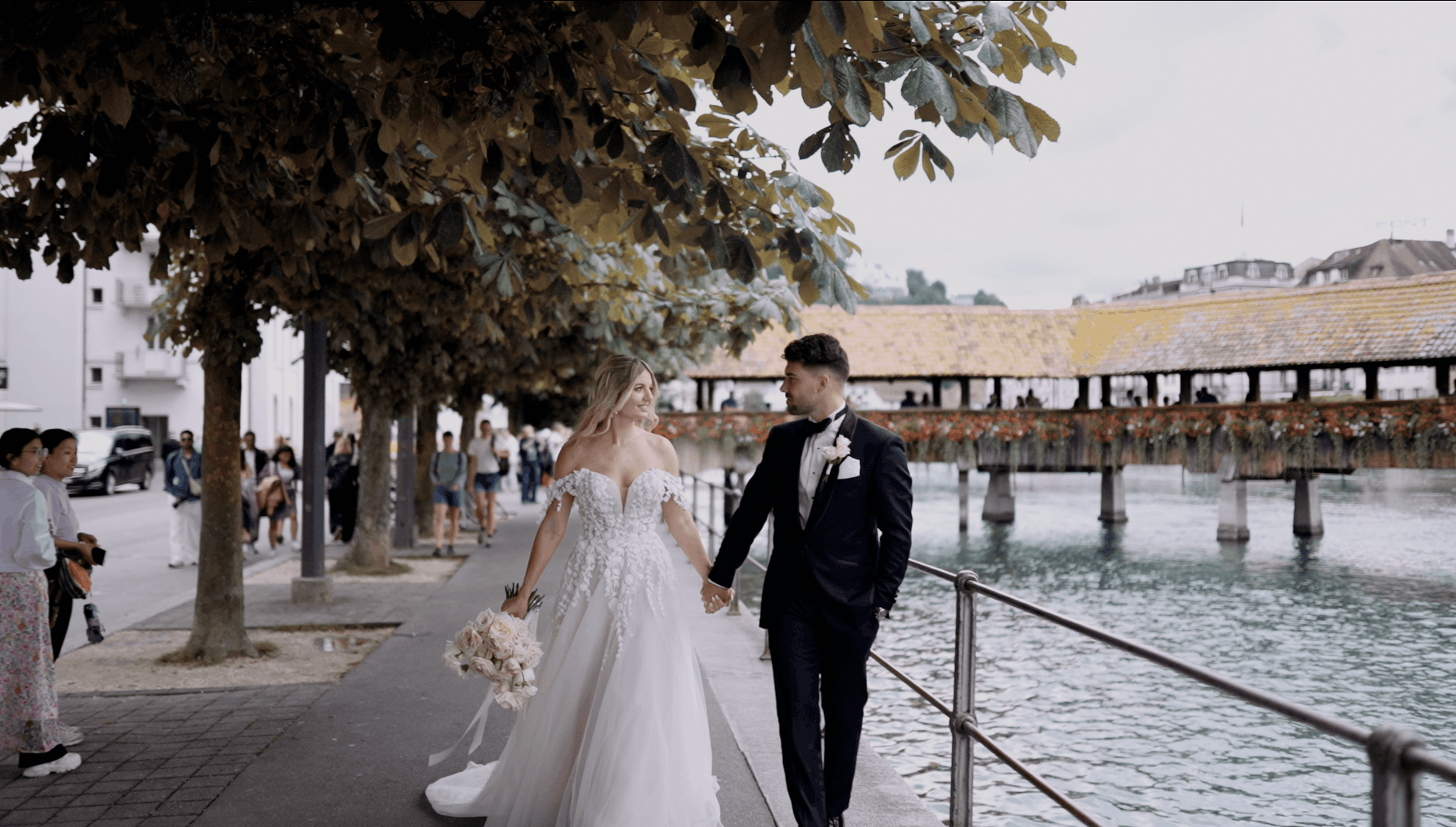 A bride and groom walking hand in hand along a waterfront promenade, surrounded by trees and onlookers, with a historic covered bridge in the background.