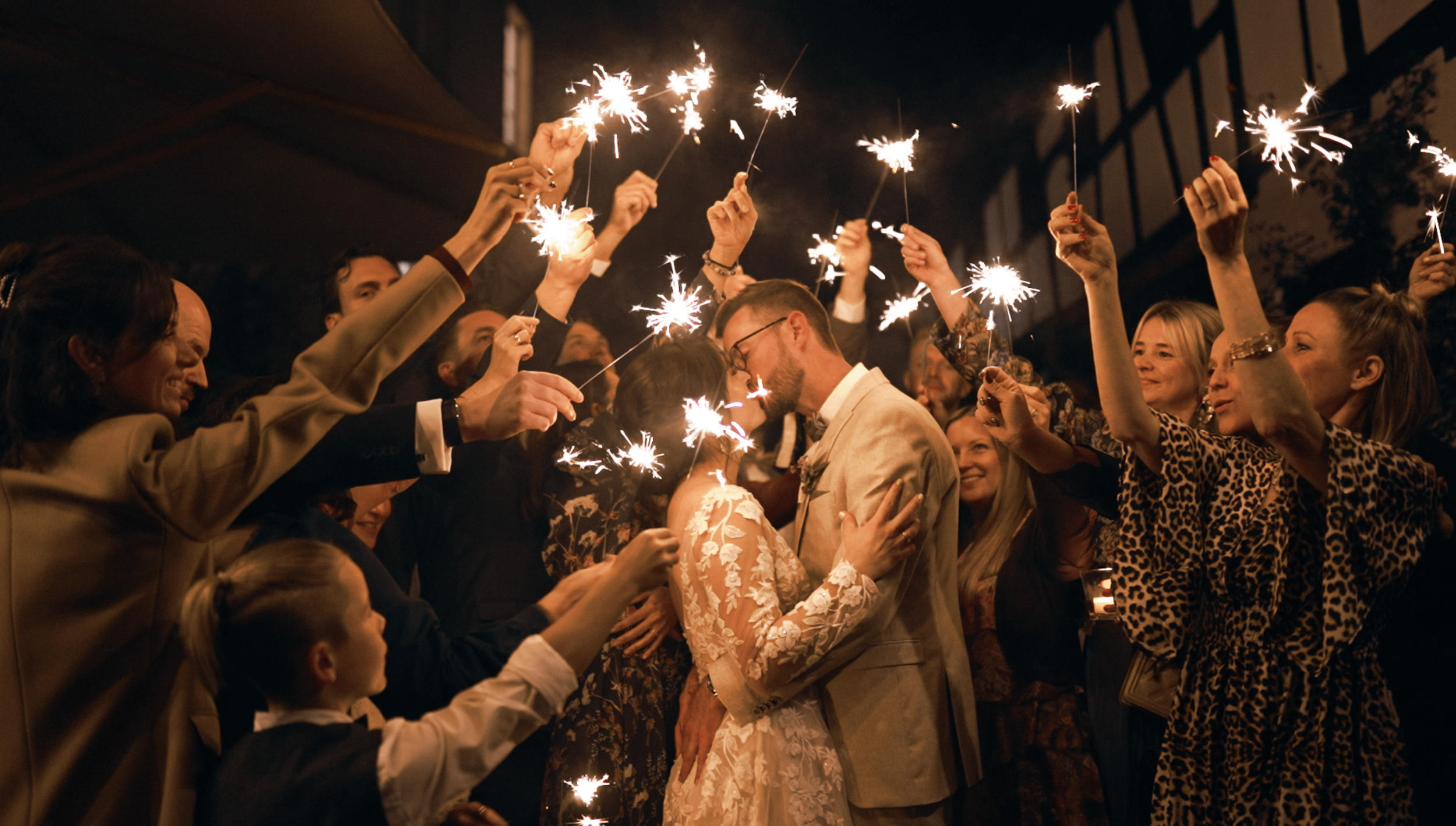 A crowd of people celebrating at night, holding sparklers and kissing, possibly a wedding or festive event.