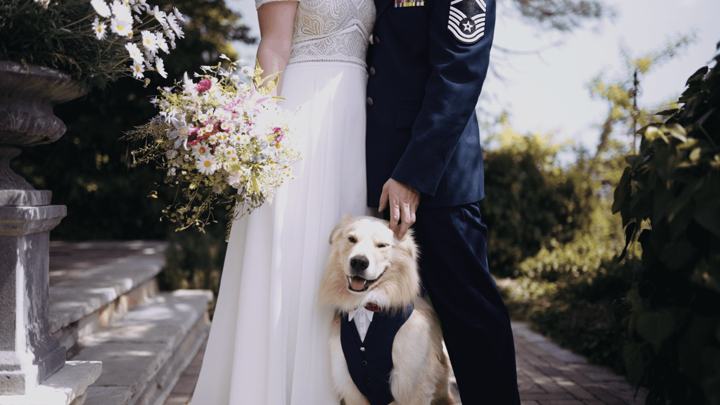 A bride and groom standing outdoors with a golden retriever dog, holding the dog's head and petting it. The bride holds a bouquet of flowers, and the groom is dressed in a military uniform, while the background shows trees and stone stairs.