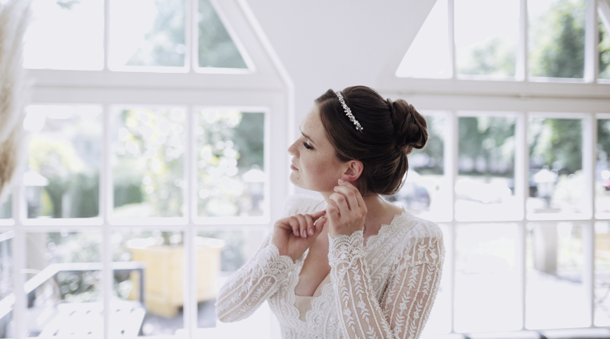 A bride with dark hair styled in an elegant updo, wearing a white lace wedding dress and a sparkling headband, adjusting an earring in front of a large window with white framing.