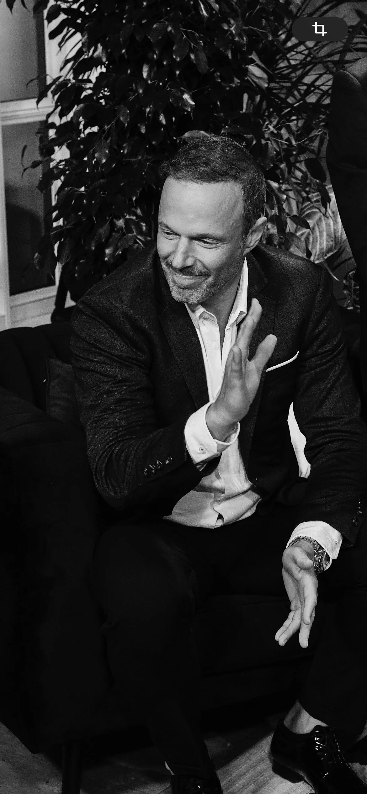 A man in a suit and white shirt sitting on a couch, smiling, with his hand raised in a greeting gesture. There are plants and a window in the background.