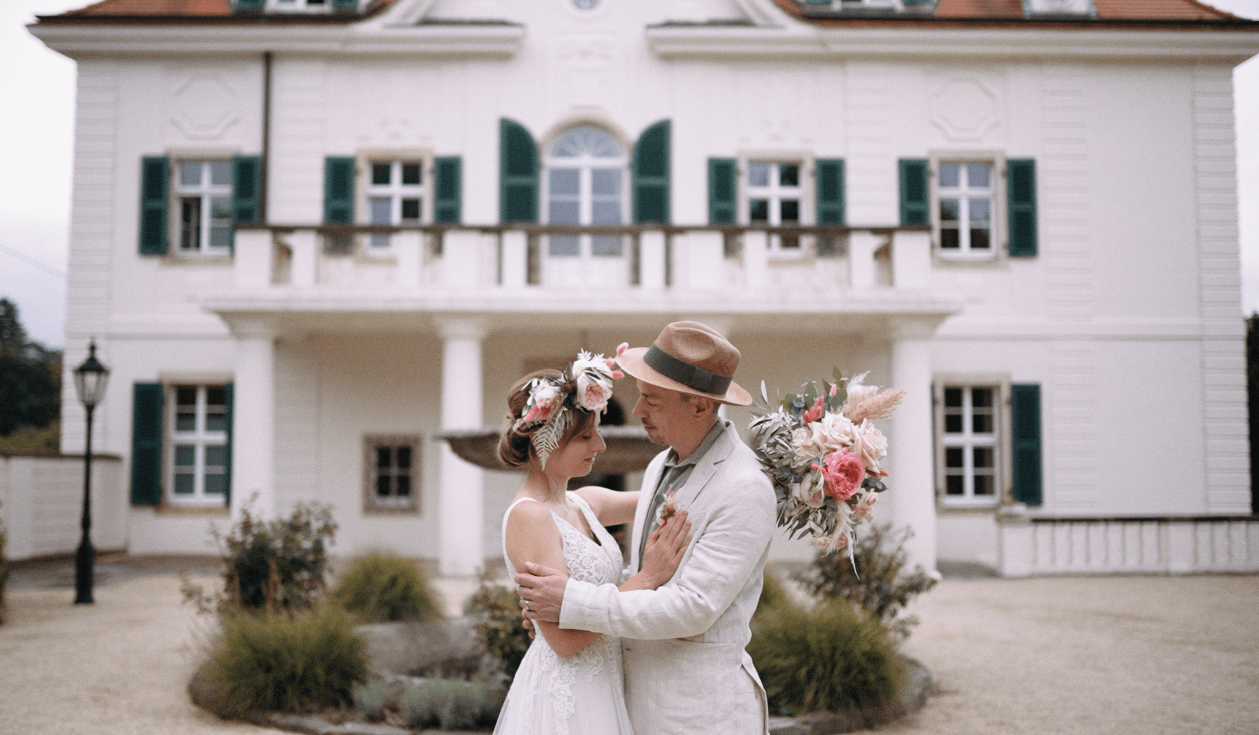 A bride and groom embrace outdoors in front of a large white house with green shutters, holding a bouquet of flowers.