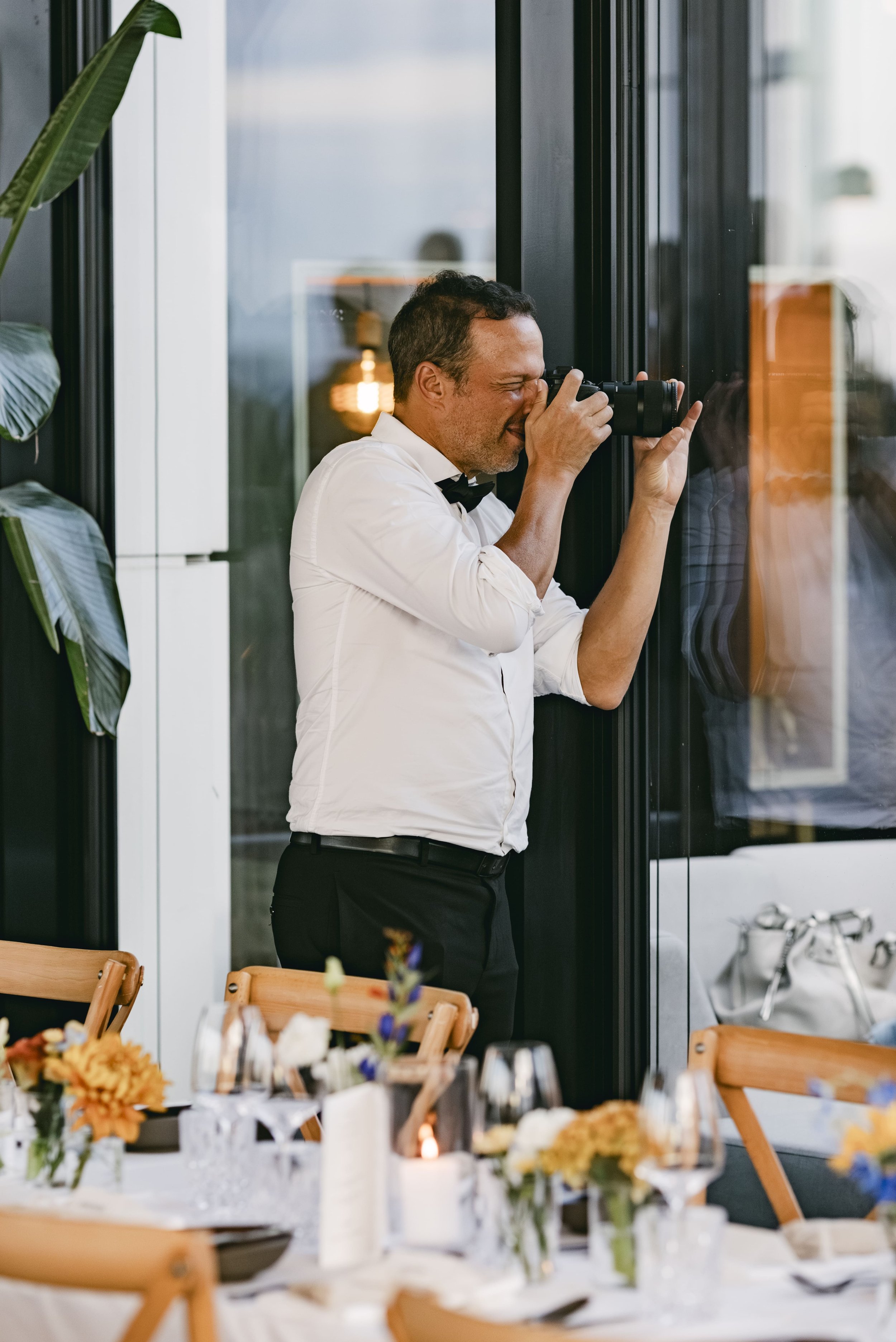 A man dressed as a waiter in a white shirt and black bow tie takes photos through a window at a dining table decorated with flowers and candles.
