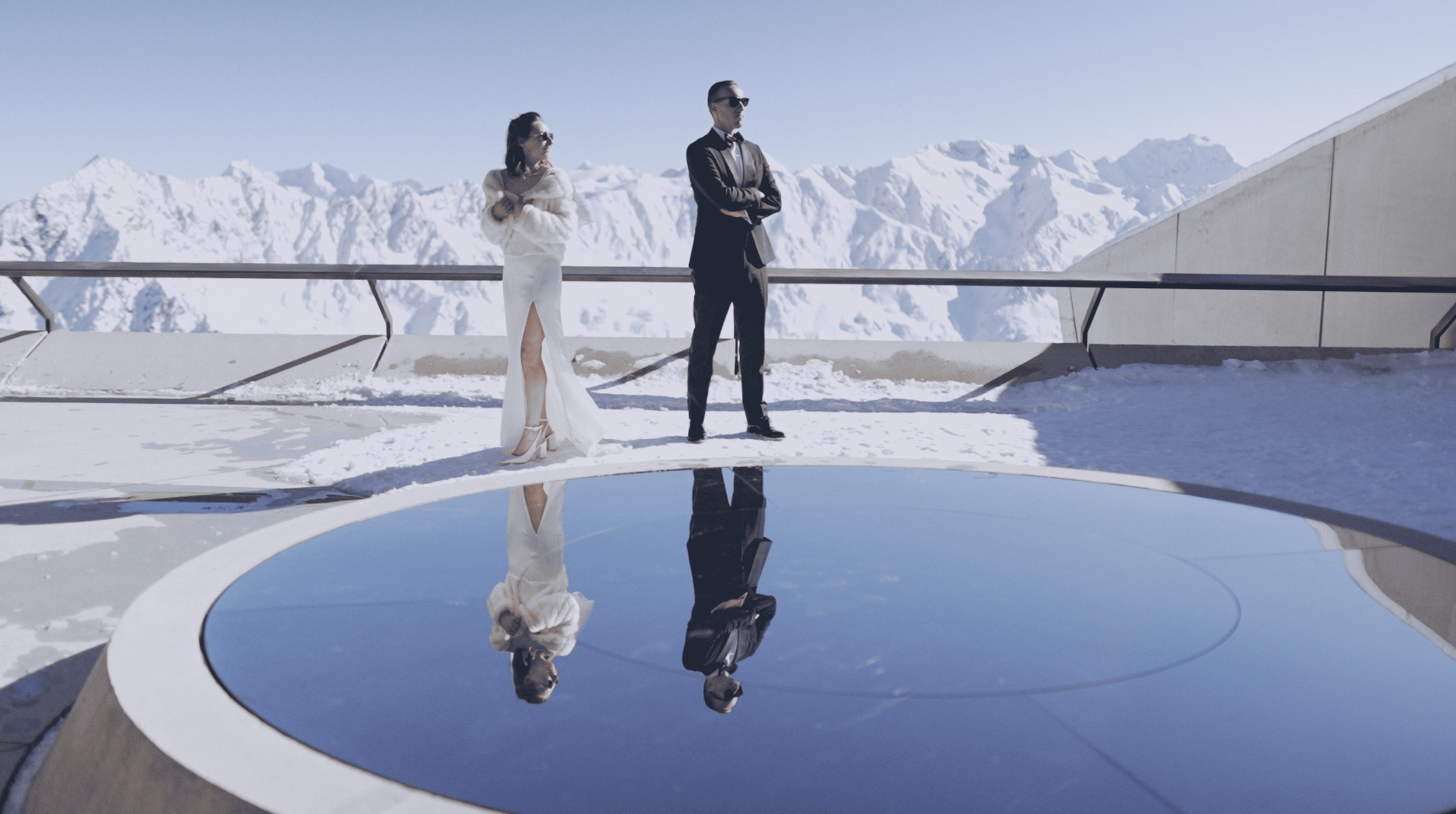 A man and woman dressed in formal attire standing by a circular water feature on a snowy mountain terrace with snow-capped peaks in the background.