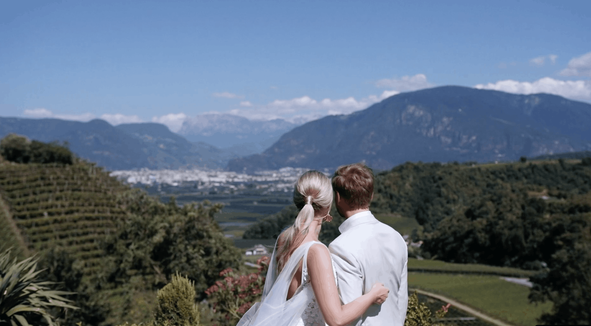 A couple with blonde hair in wedding attire stands close together overlooking a scenic valley with green hills and mountains, under a blue sky.