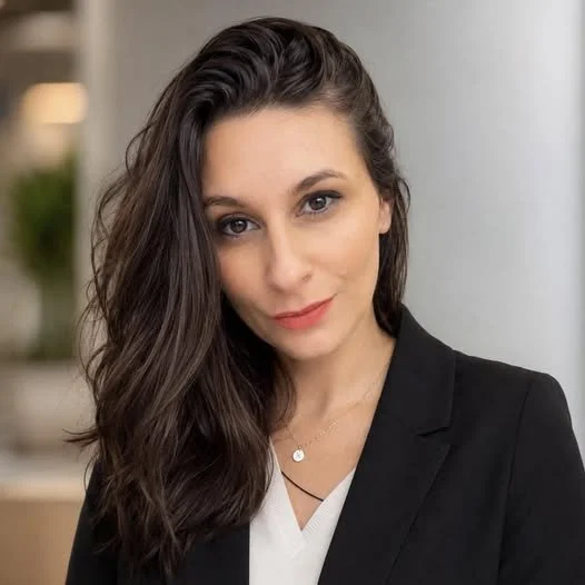 A woman with long dark hair and light makeup, wearing a black blazer over a white top, looking at the camera with a slight smile, in an indoor setting with blurred background.