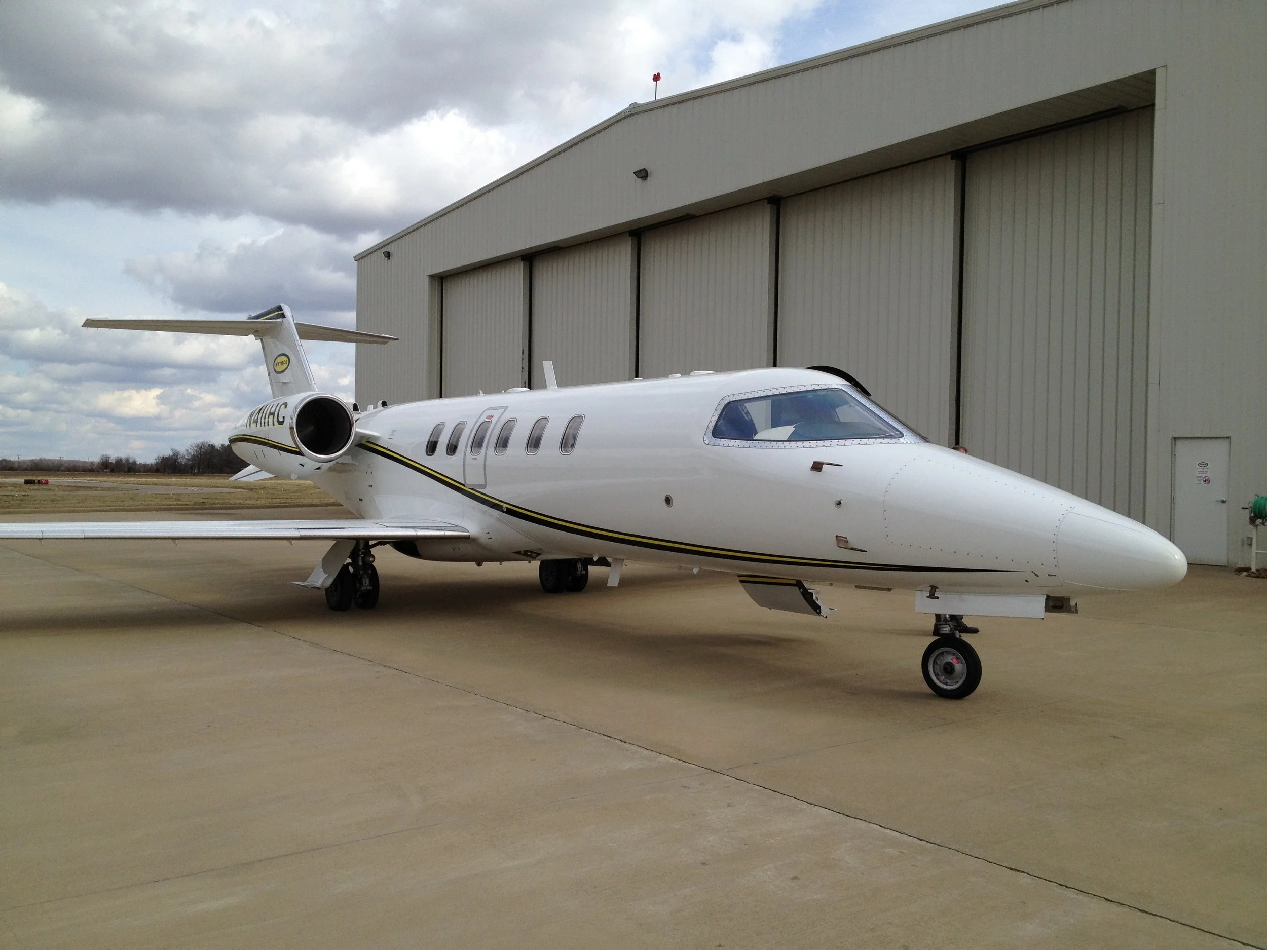 White private jet parked on the tarmac next to a large beige hangar under a cloudy sky.