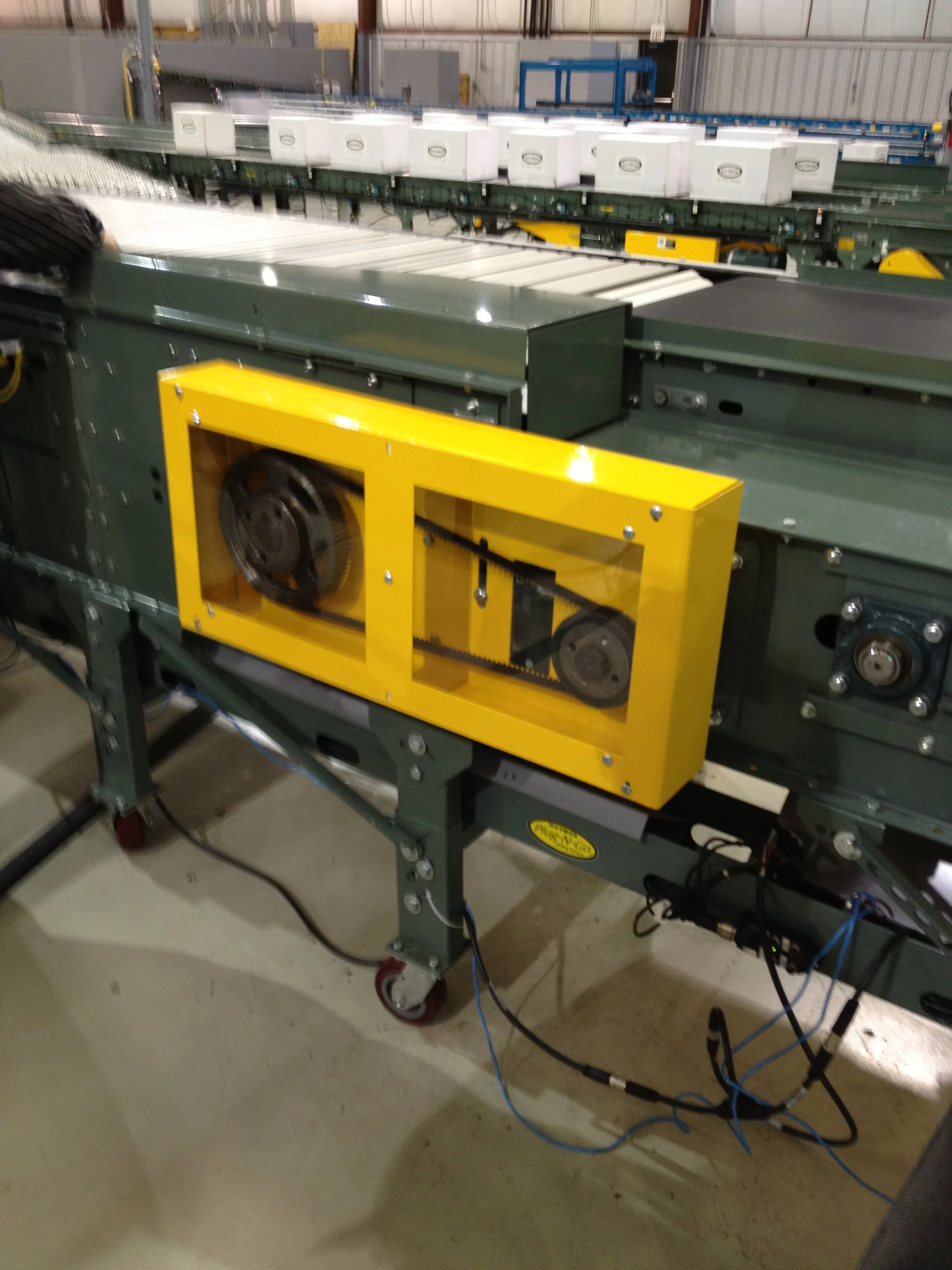 Industrial conveyor system with white boxes on a green frame in a warehouse or factory setting.