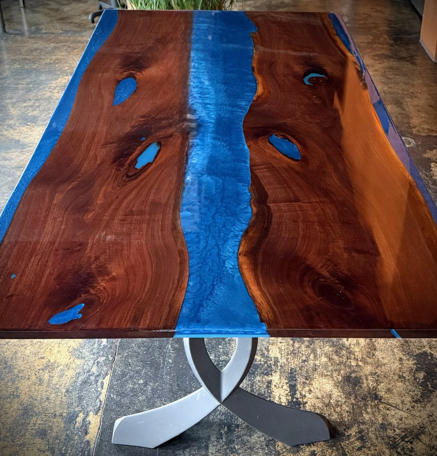 A wooden dining table with a live edge design, featuring a central blue resin river running through it, and an ornate white metal leg base.