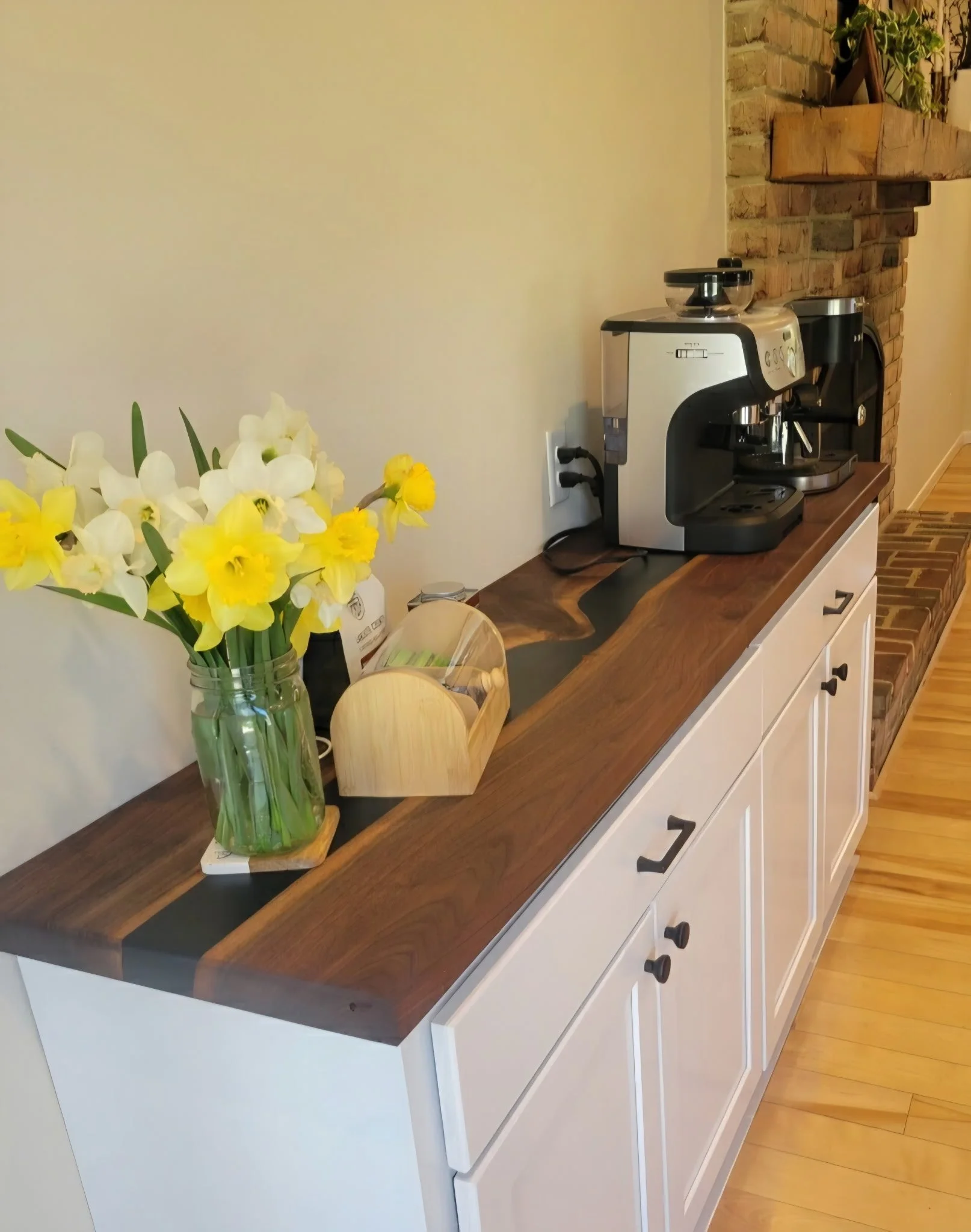 A kitchen counter with a bouquet of yellow and white flowers in a glass vase, a coffee machine, and a wooden tray, against a white wall and brick backsplash.