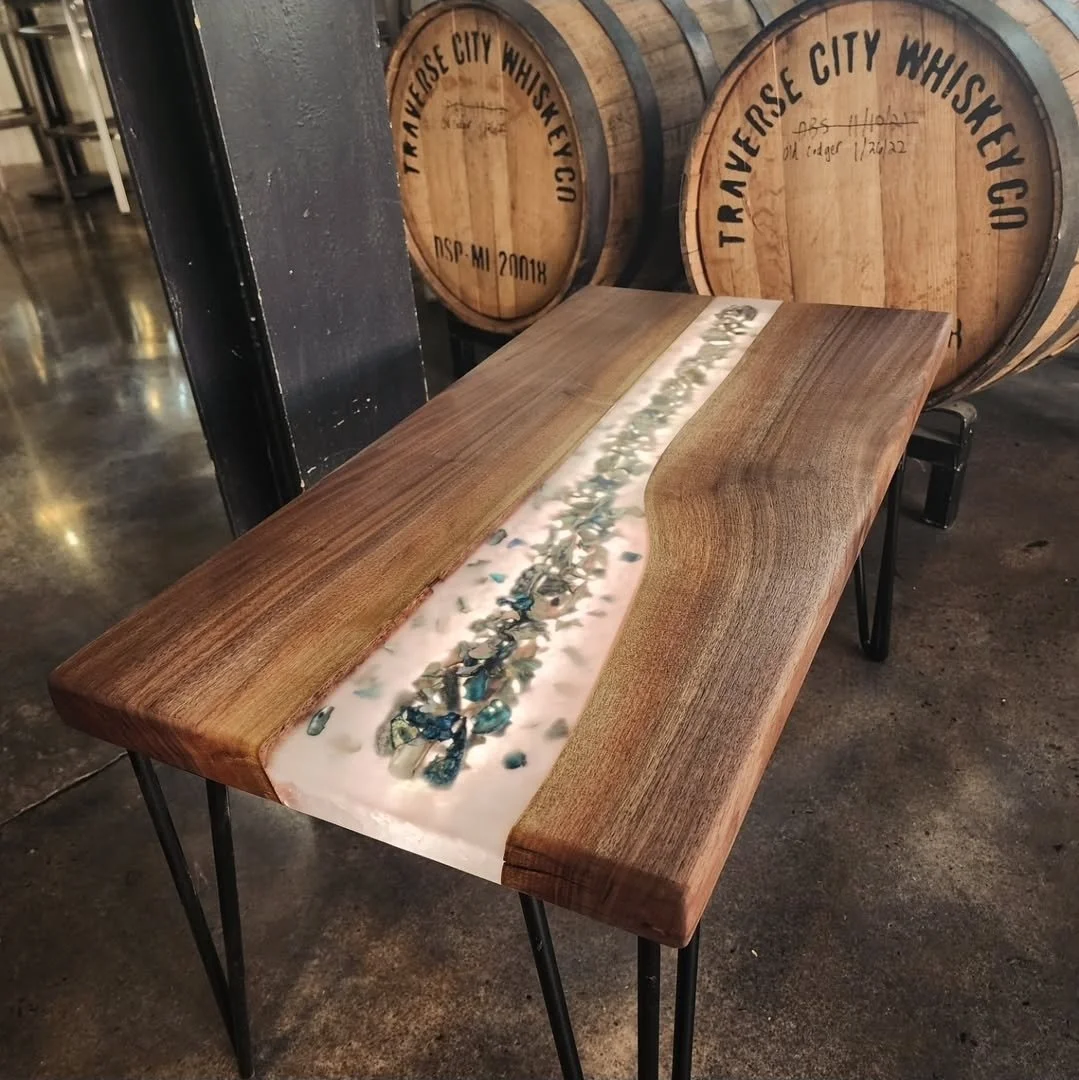 A wooden table with a resin river running through the center, embedded with stones. In the background, there are whiskey barrels labeled "Traverse City Whiskey Co."
