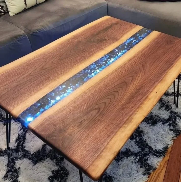 Wooden table with a glowing resin inlay running through the center, set in a living room with a gray sofa and a patterned rug.