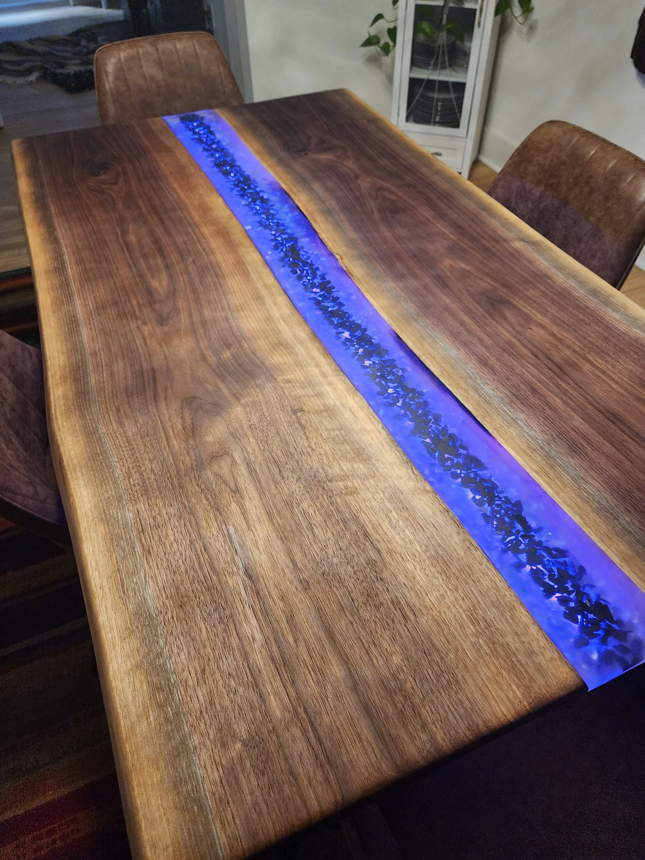 A wooden dining table with a blue backlit resin inlay and brown leather chairs in a dining room.