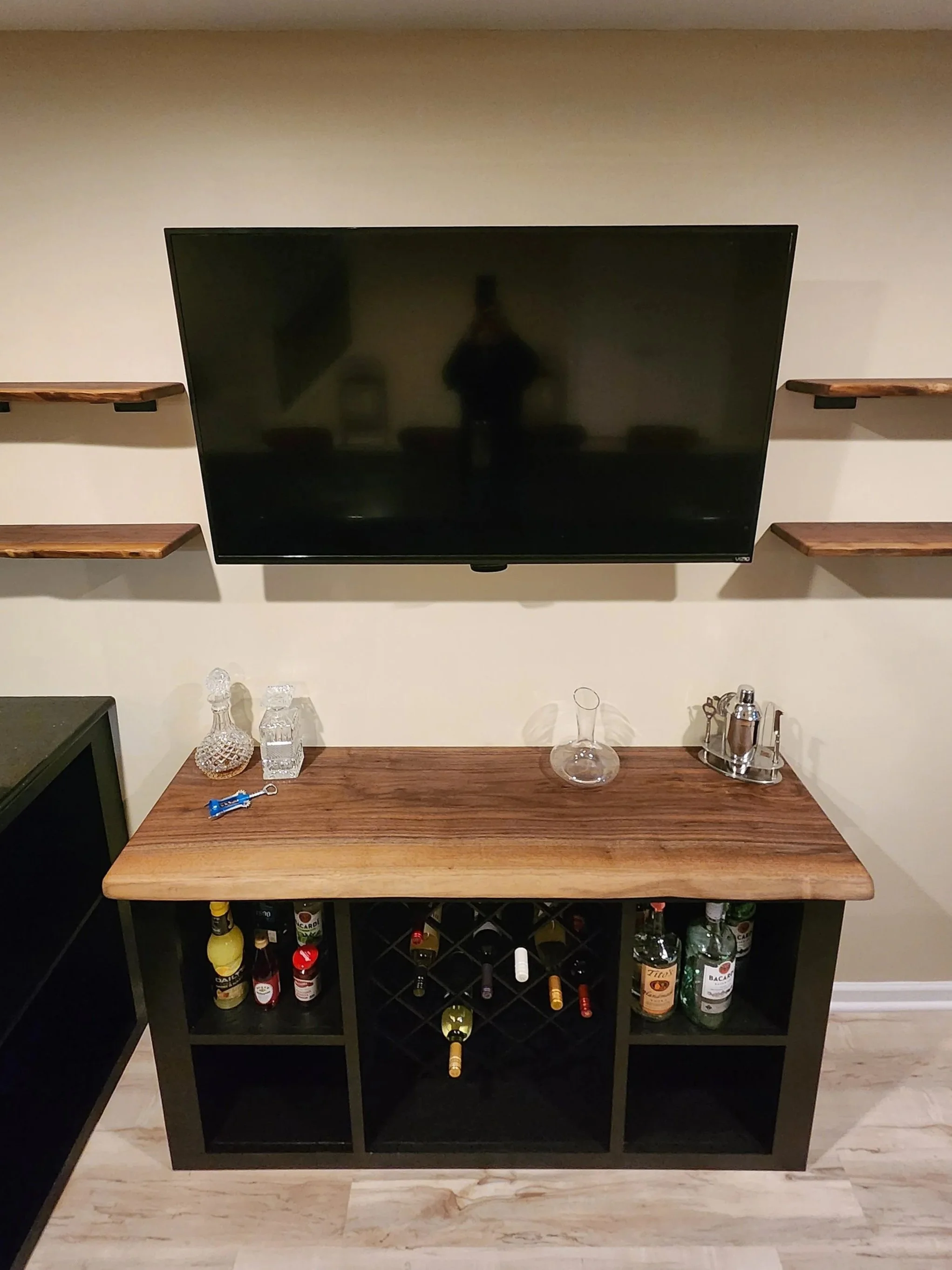 A black wine rack with bottles of alcohol inside, placed against a wall. On top of the rack, there is a small wooden table with glass decanters, water bottles, and a cocktail shaker. Above the table, a flat-screen TV is mounted on the wall, and there are two small wooden shelves on either side of the TV.