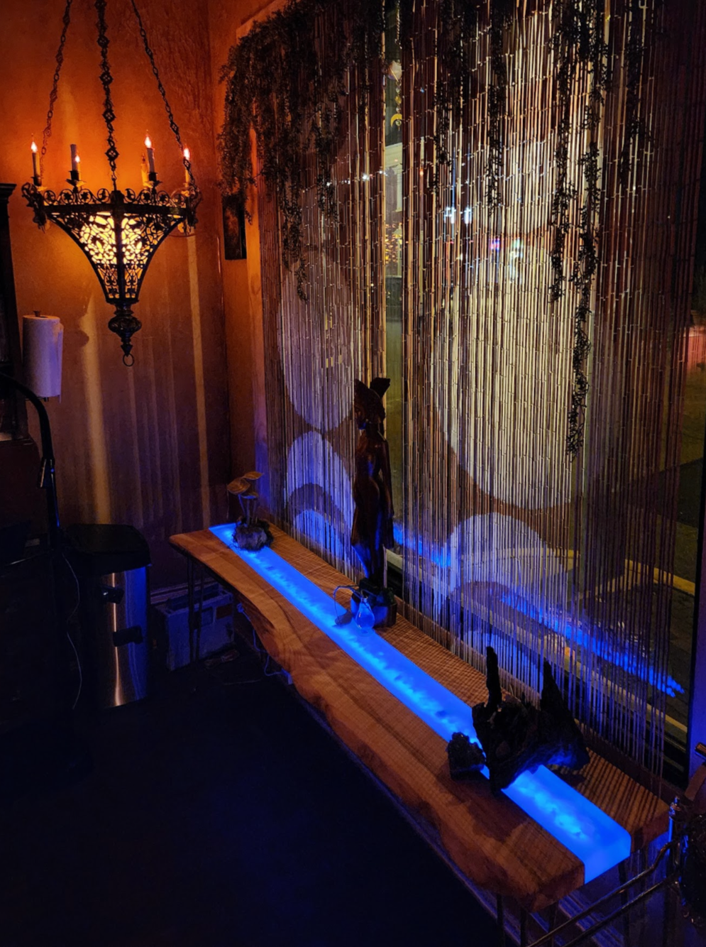 Interior of a restaurant or bar with dim lighting, a large decorative chandelier, a bamboo curtain, sculptures, and blue LED lighting.