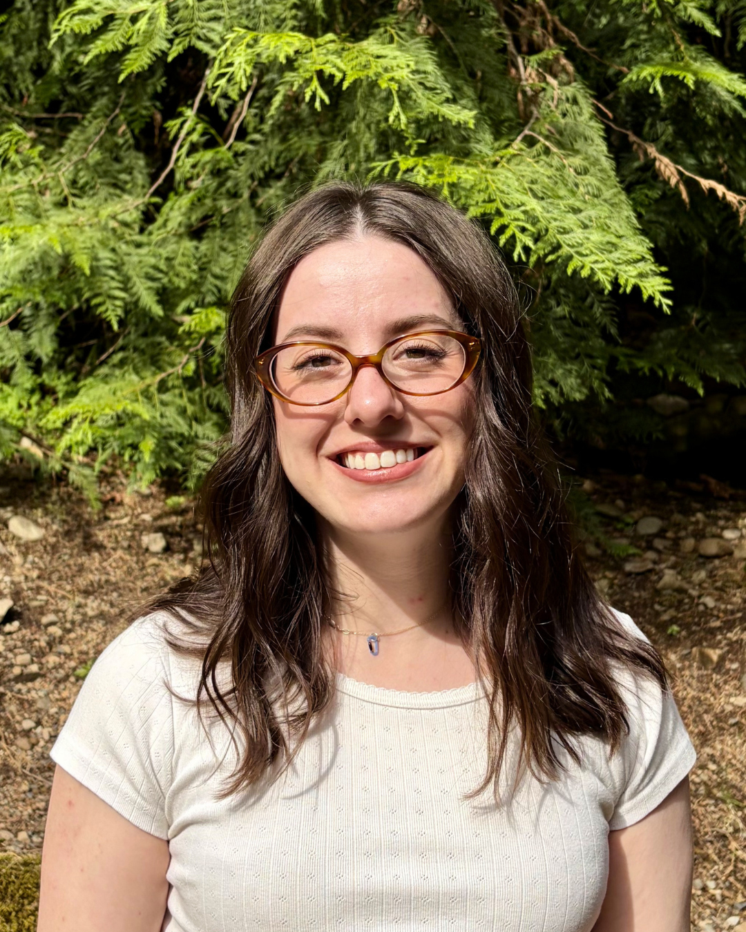 A young woman with brown hair, wearing glasses and a white shirt, smiling outdoors with green foliage behind her.