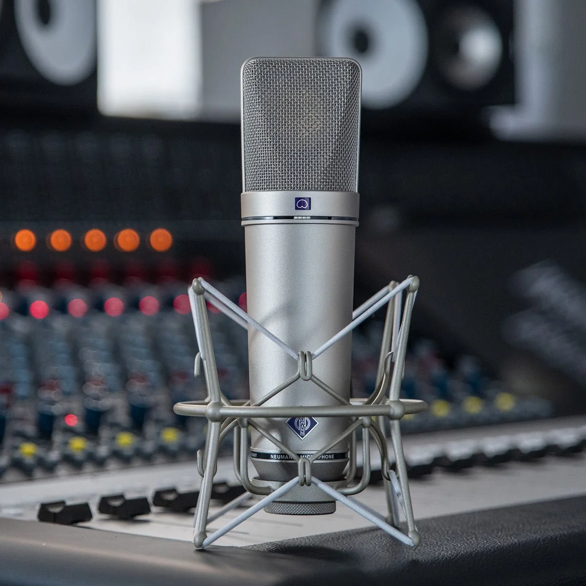 A large studio condenser microphone with a silver finish positioned in front of an audio mixing console, surrounded by a shock mount.