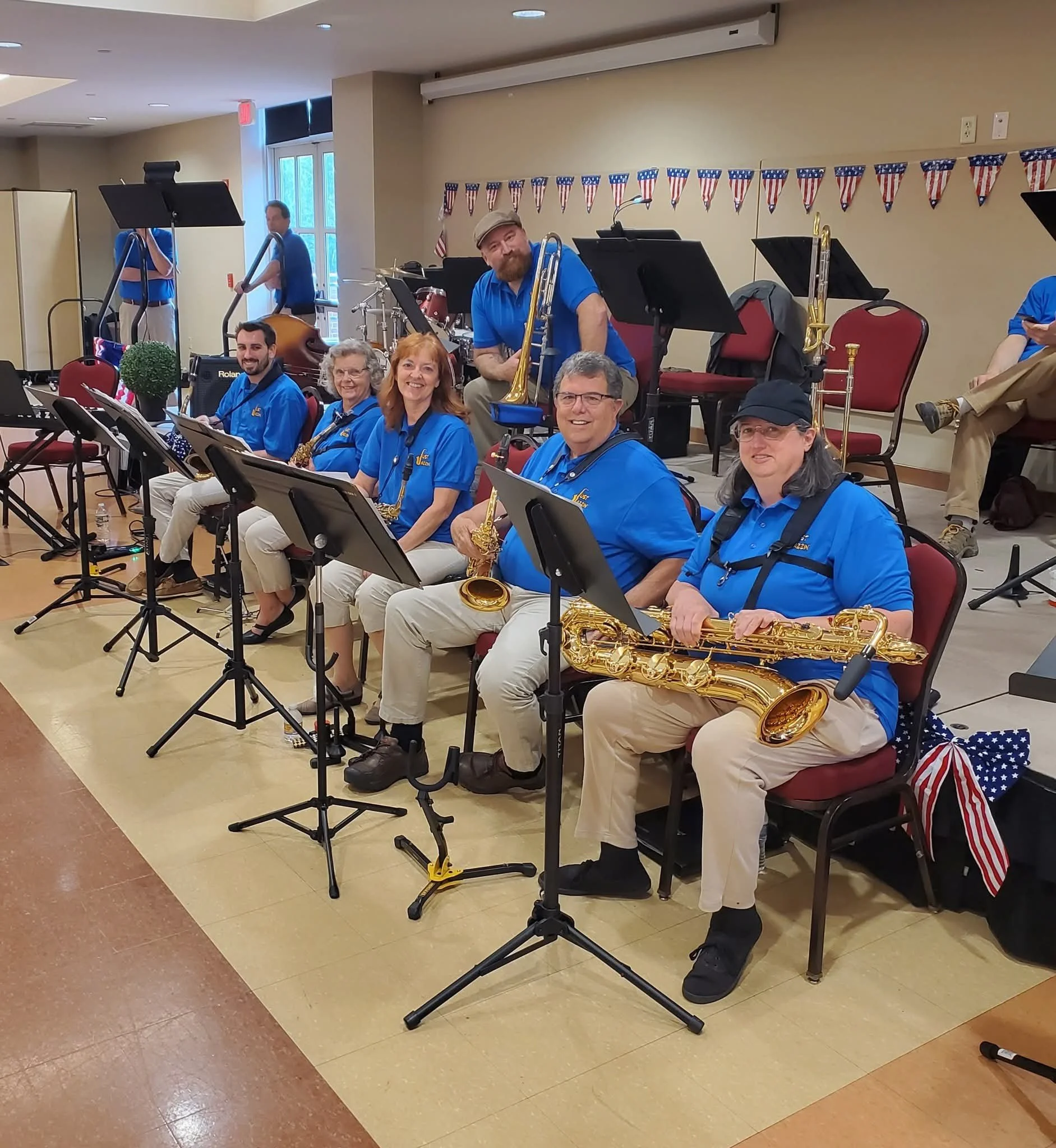 A group of musicians in blue shirts, sitting in a line, holding saxophones, in a room decorated with red, white, and blue patriotic banners.