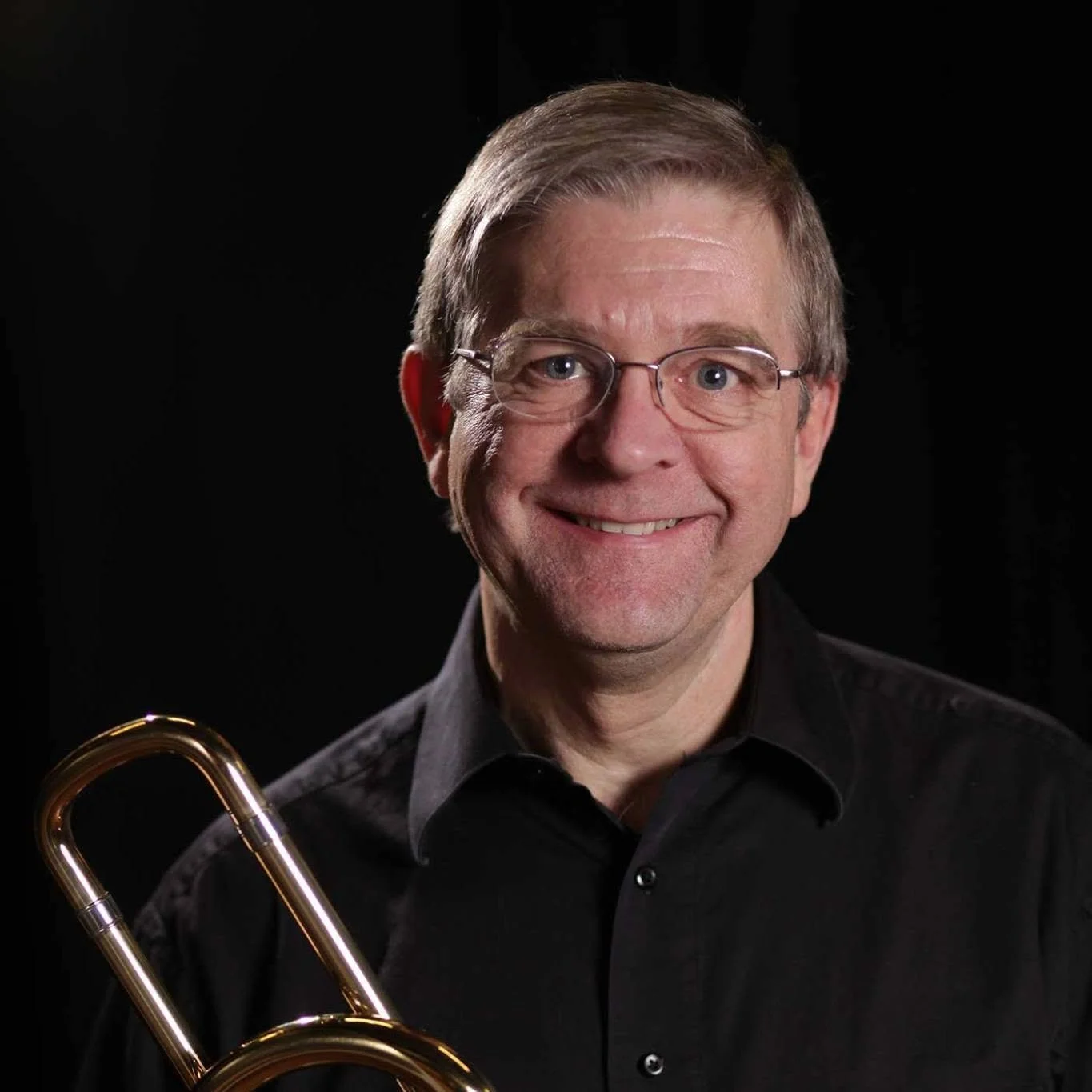 A smiling man with glasses, light brown hair, and a black shirt holding a brass trombone against a black background.