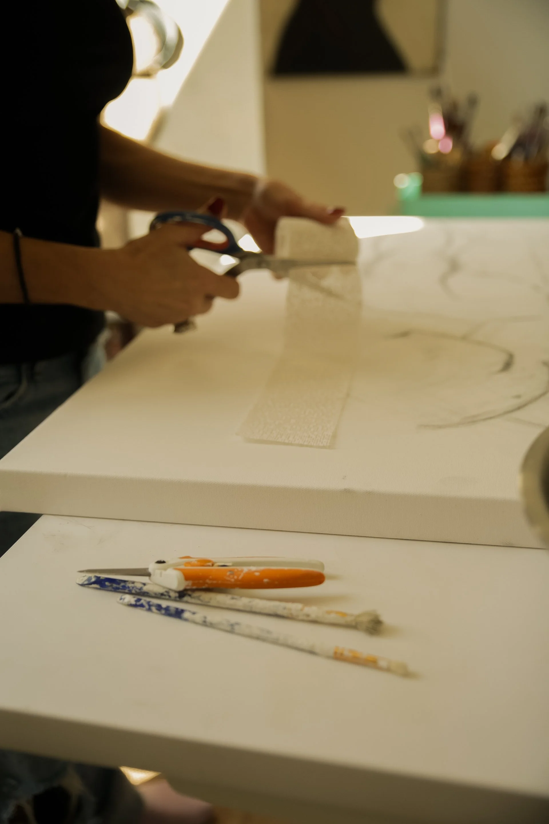 Person cutting white fabric with scissors at a worktable, with paintbrushes in the foreground