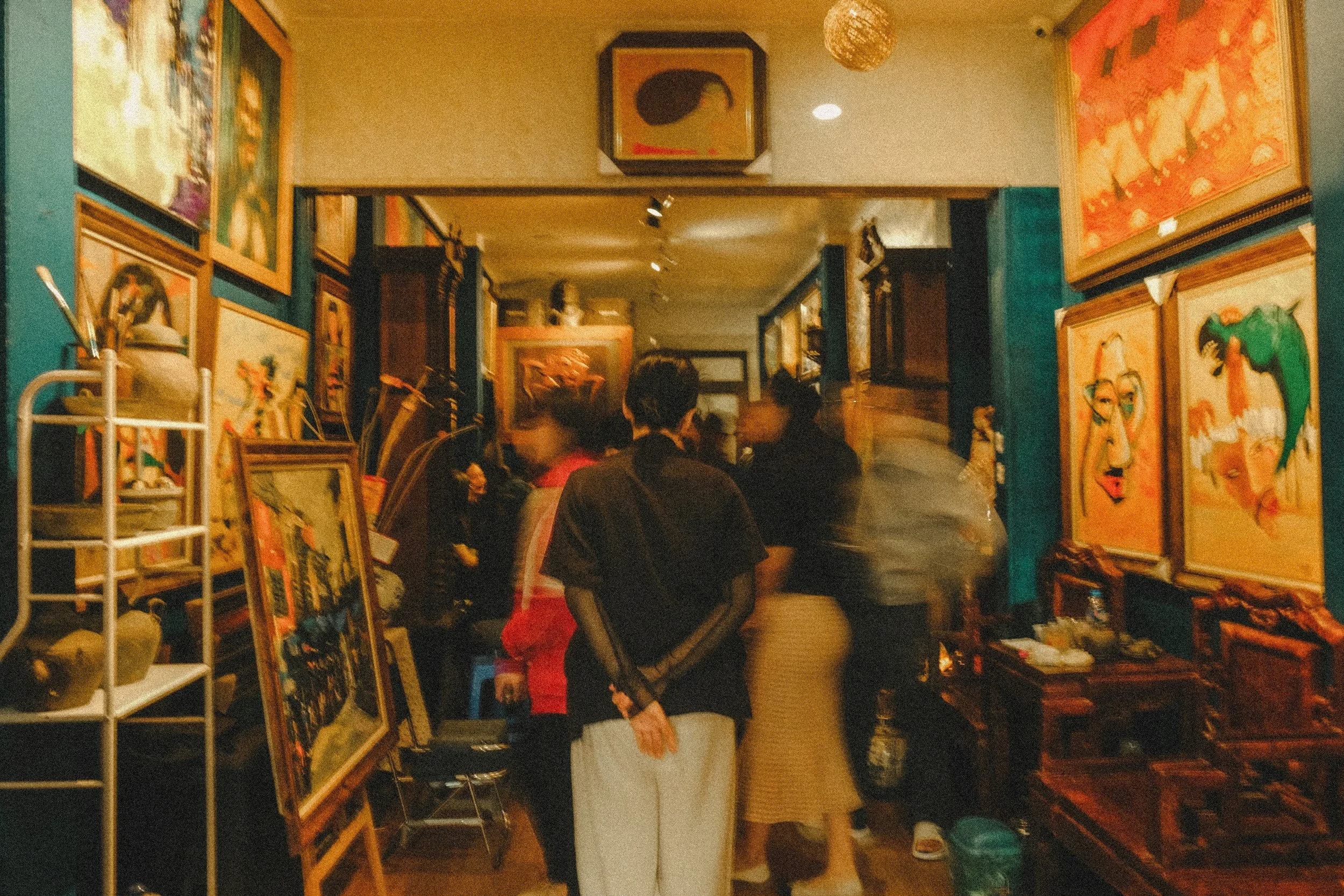 Interior of an art gallery or studio with various framed paintings on the walls and displayed on easels. Several people are observing the artwork, and there are shelves and other furniture inside.