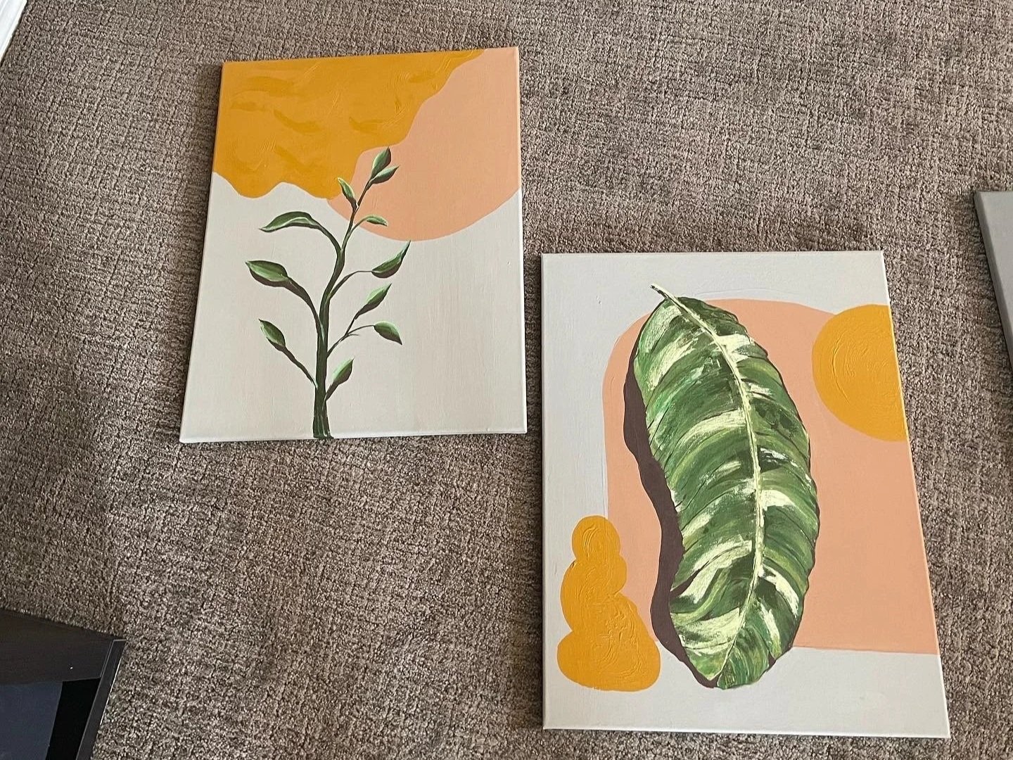 Two paintings on canvas depiciting abstract botanical themes with green leaves and yellow-orange shapes, placed on a textured brown carpet.
