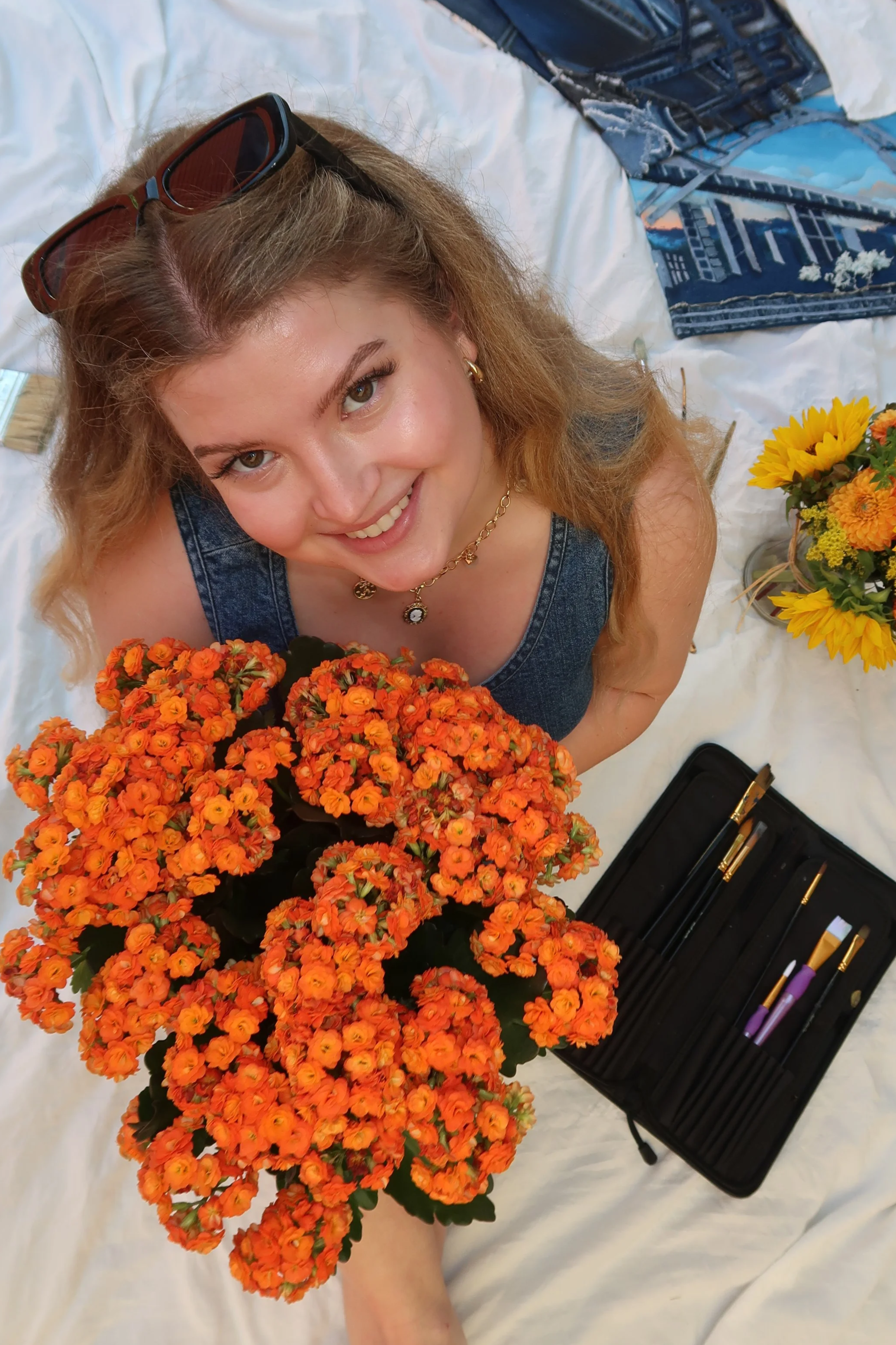 A woman with wavy blonde hair, wearing sunglasses on her head, smiling at the camera while sitting on a white surface. She is surrounded by orange flowers and has a bouquet of sunflowers nearby. There are paintbrushes and other painting tools in a black case beside her.