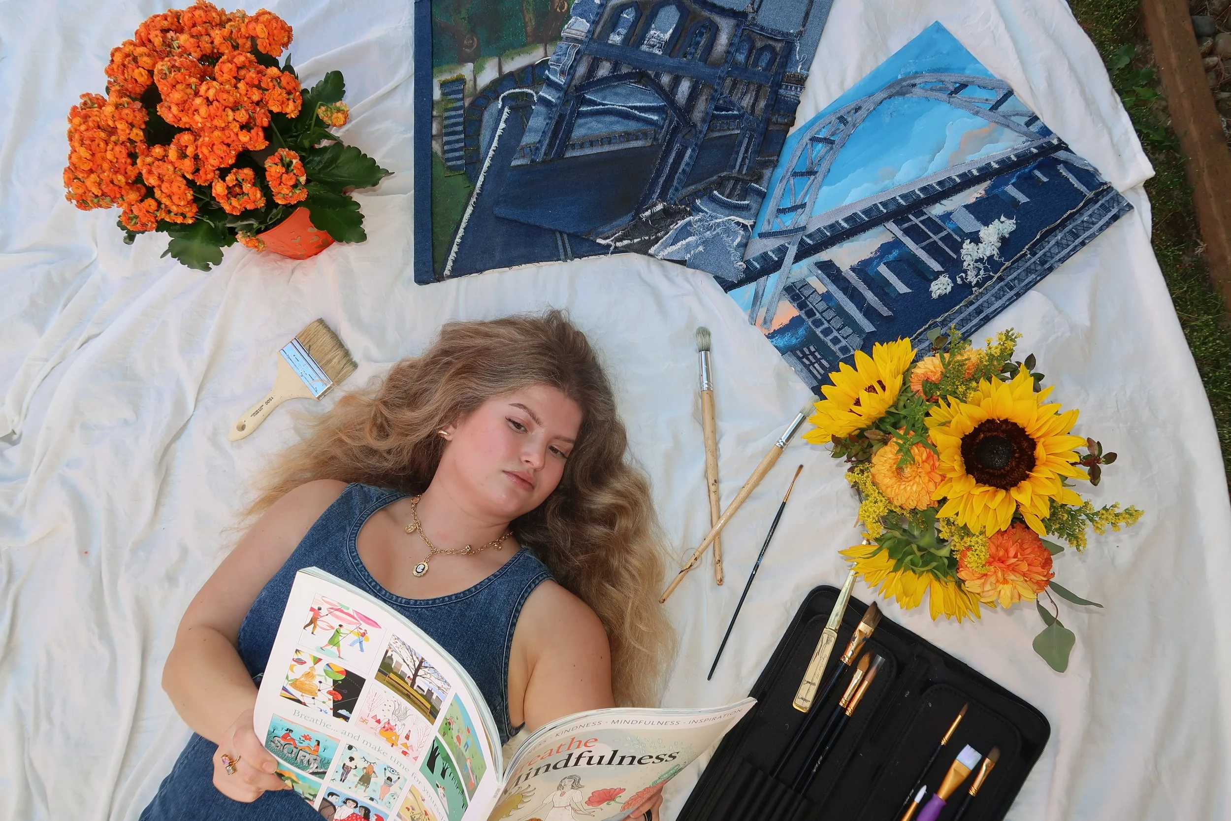 A young woman lying on a white cloth in a park or garden setting, surrounded by colorful flowers including orange and yellow blooms, reading a book about mindfulness and meditation. Nearby are paint brushes, a paint palette, and a box of tools for painting, with unopened paintings of city bridges and a natural landscape.