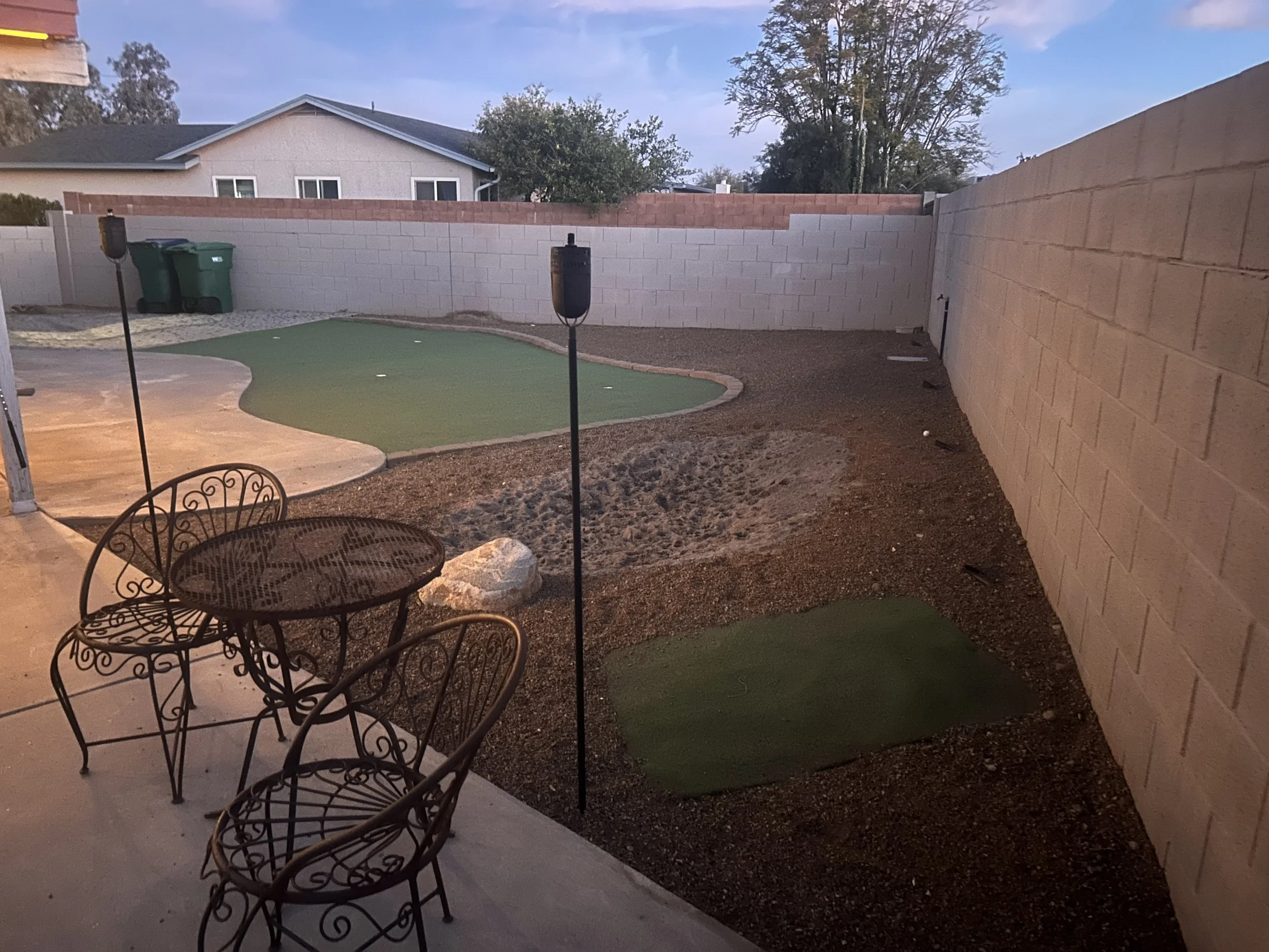 Backyard patio with metal chairs, a small putting green, mulch ground, a wall, and trees in the background during dusk.