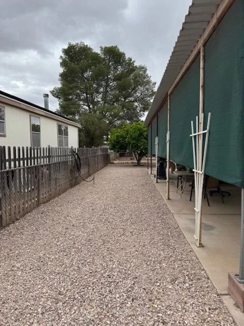 Gravel pathway between a fence and a building with a green canopy, tables, and chairs on the right, and a tree at the end of the pathway.