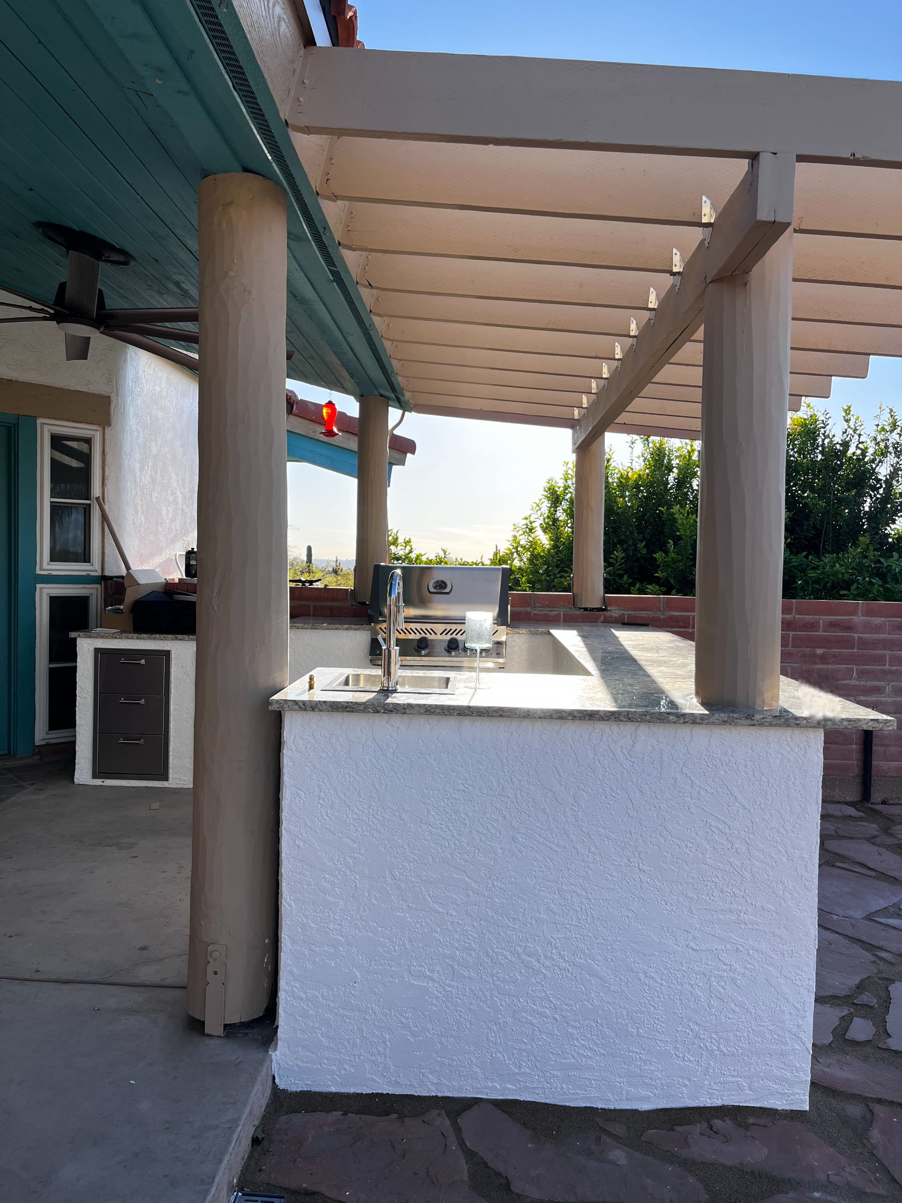 An outdoor kitchen area with a granite countertop and a built-in grill, covered by a wooden pergola with white beams, against a backdrop of greenery and a clear blue sky.