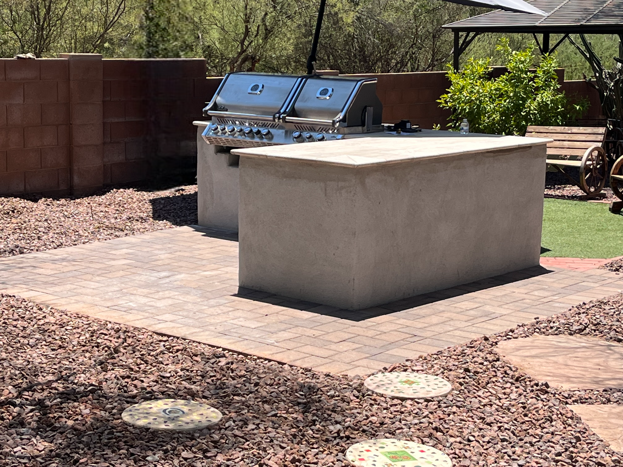An outdoor kitchen area with a modern stainless steel barbecue grill on a white stone counter, set against a brick wall with trees and shrubs in the background, and a paved patio with decorative stepping stones.