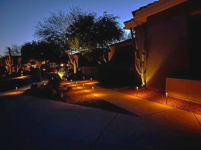 Neighboring houses illuminated by outdoor pathway lights at dusk, with trees and clear sky in the background.