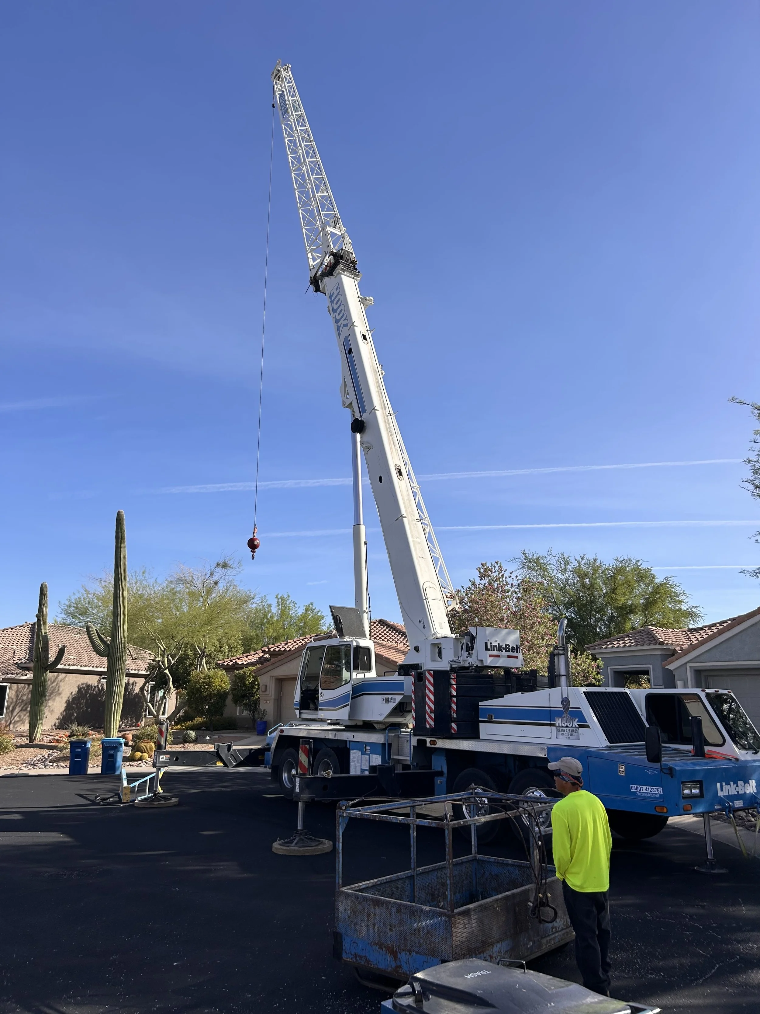A crane lifting a heavy object in a residential neighborhood with single-story houses, trees, and a clear blue sky.