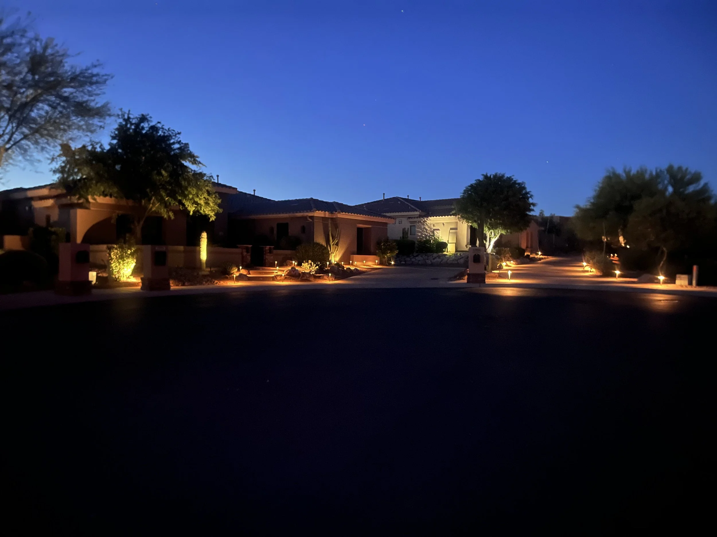 Nighttime scene of a residential neighborhood with dark sky, trees, and houses illuminated by outdoor lighting.