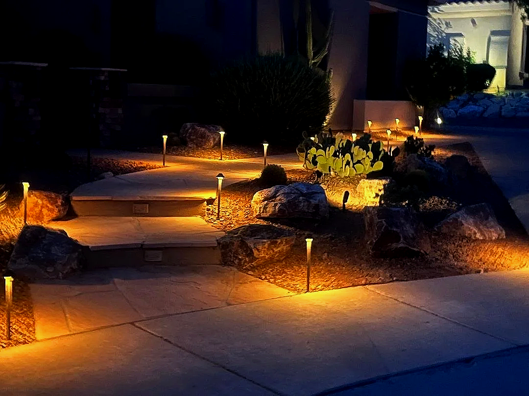 Nighttime view of a landscaped front yard illuminated by pathway lights, with rocks, bushes, and cactus plants.