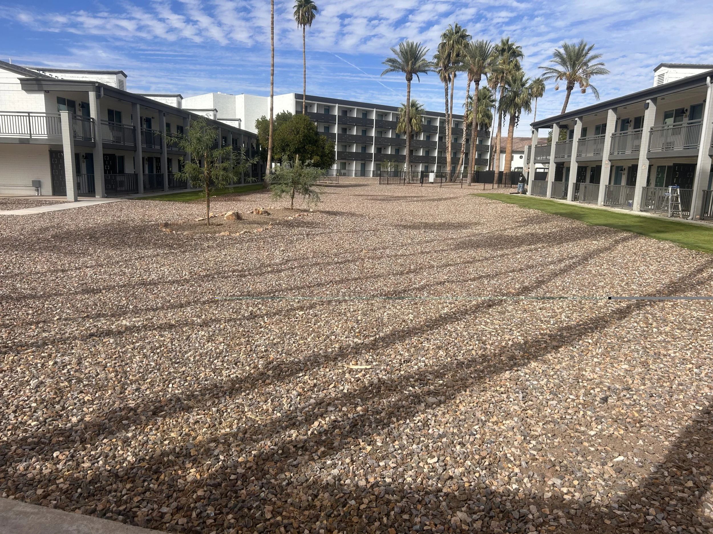 View of a modern apartment complex with three buildings, tall palm trees, a cleared gravel area, and a partly cloudy sky.