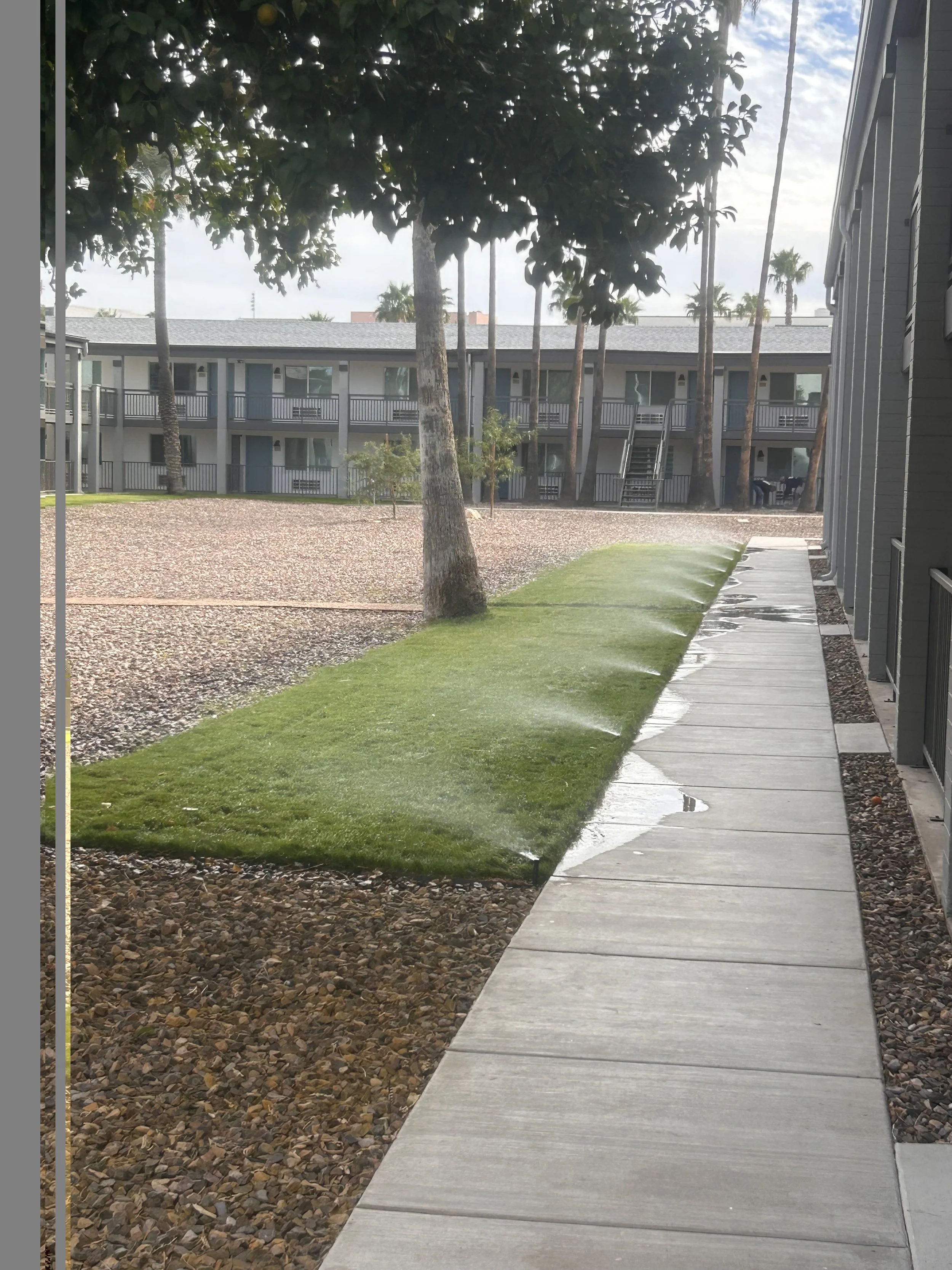 Sidewalk with grass and gravel area, apartment building with balconies, trees, and a cloudy sky in the background.