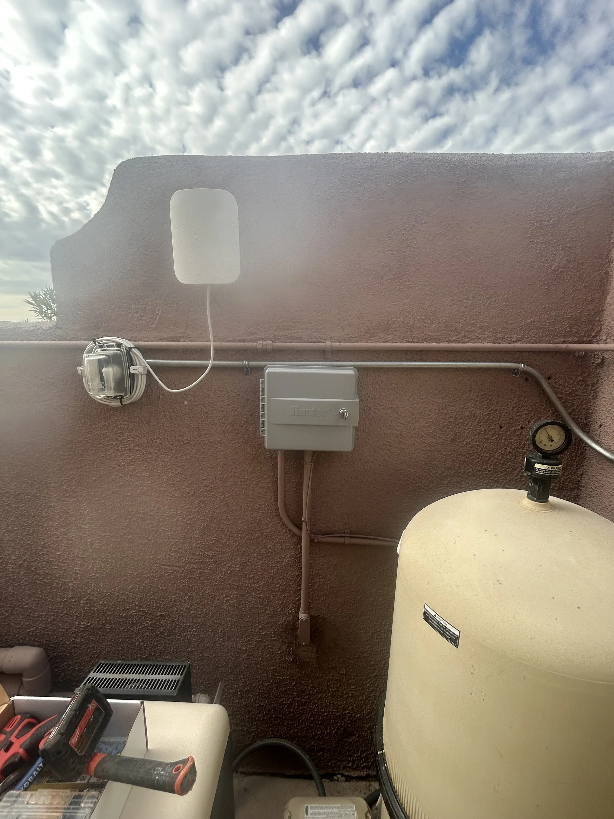An outdoor wall with a plastic electrical box, a mounted small white device, a large beige water heater, and miscellaneous tools on a nearby table.