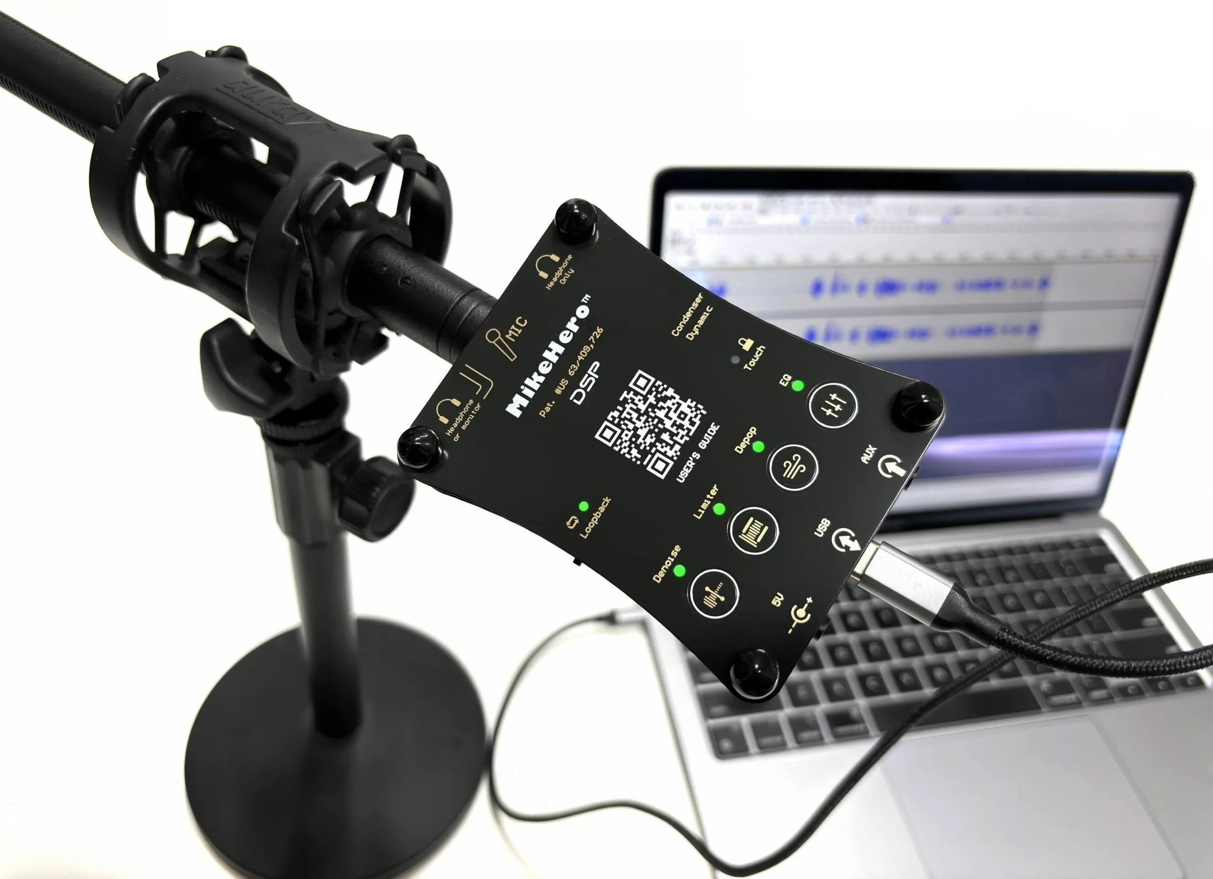 Close-up of an audio recording setup featuring a microphone mounted on a stand with a black windscreen, a digital audio interface or control panel with various buttons, a QR code, and a laptop with an open digital audio workstation software screen in the background.