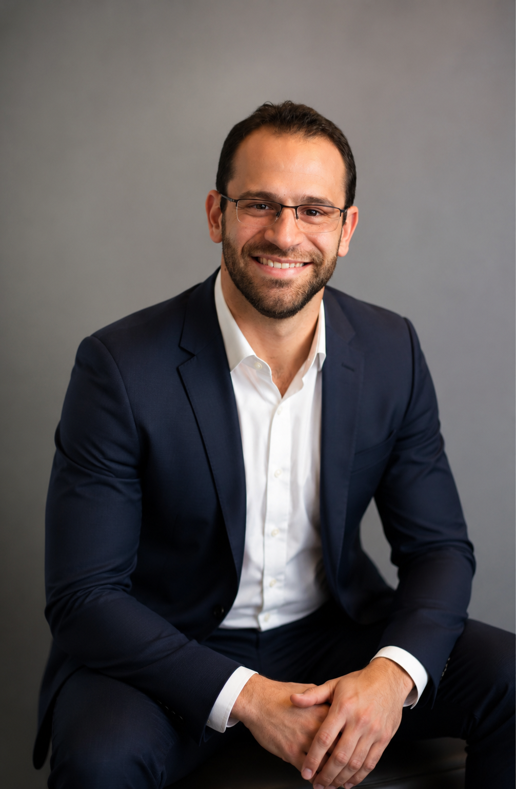 Professional man smiling, wearing glasses, a dark blue suit, and a white shirt, sitting against a gray background.