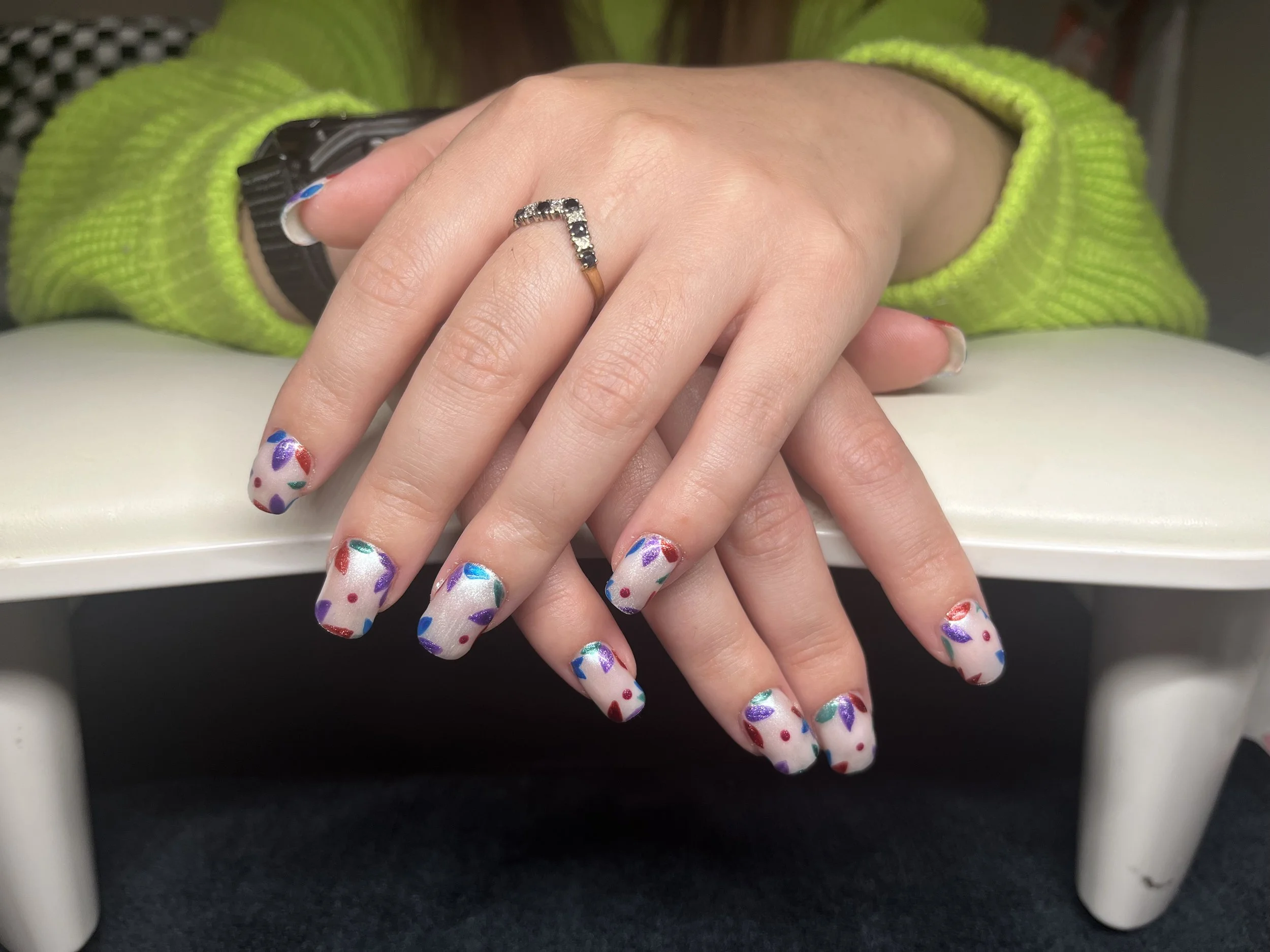 Close-up of a hand with colorful rainbow speckled nail polish, resting on top of another hand wearing a ring with black and clear stones, against a lime green sleeve background.