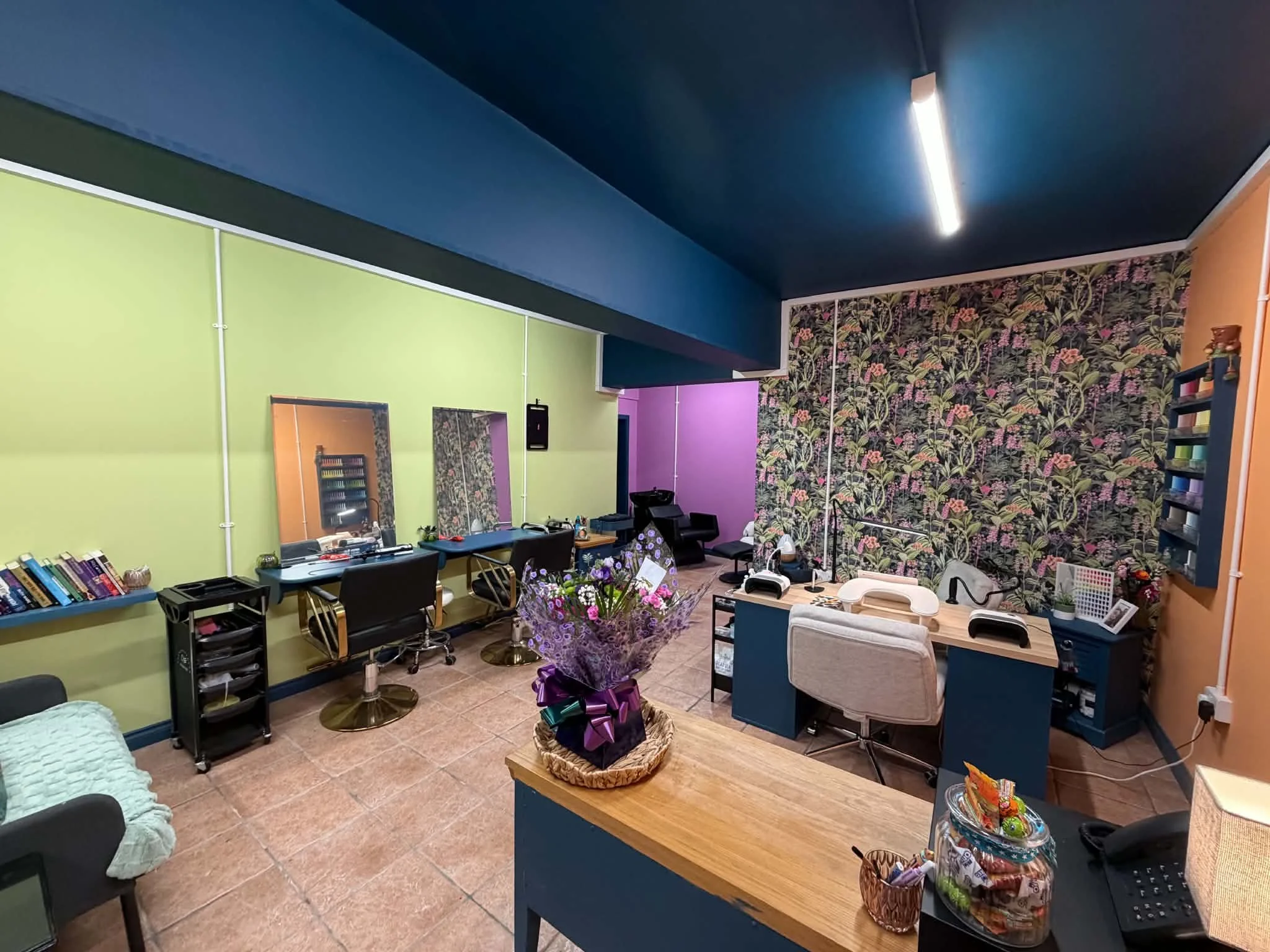 A colorful interior room with green, purple, and floral wallpapered walls, featuring multiple chairs, a desk with mirrors, books, plants, and a collection of items on a counter and shelves, with a ceiling light and tiled floor.