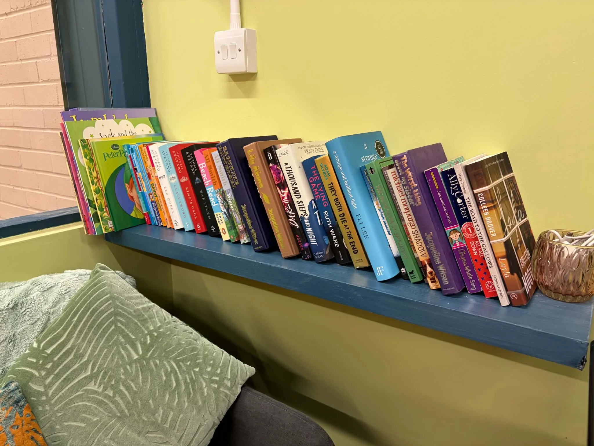 A row of colorful children's books arranged on a blue shelf against a green wall, with a green pillow with a leaf pattern on a chair in the foreground.