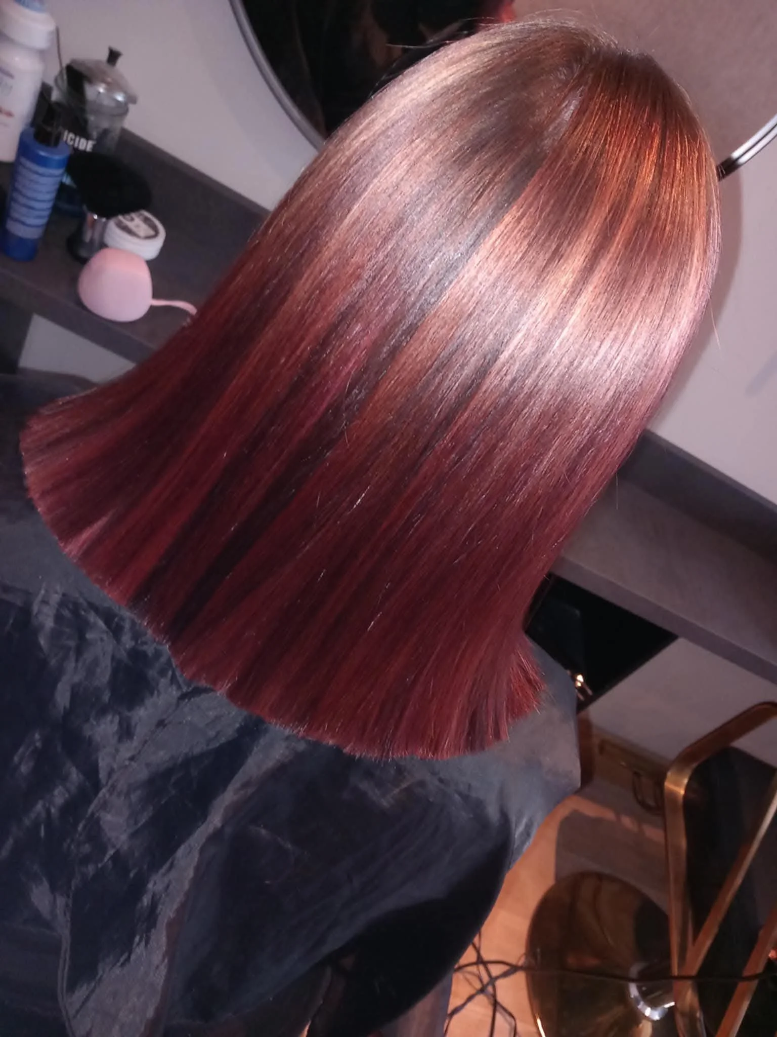 Long, straight, shiny, reddish-brown hair on a person's head, with a mirror and styling products in the background.