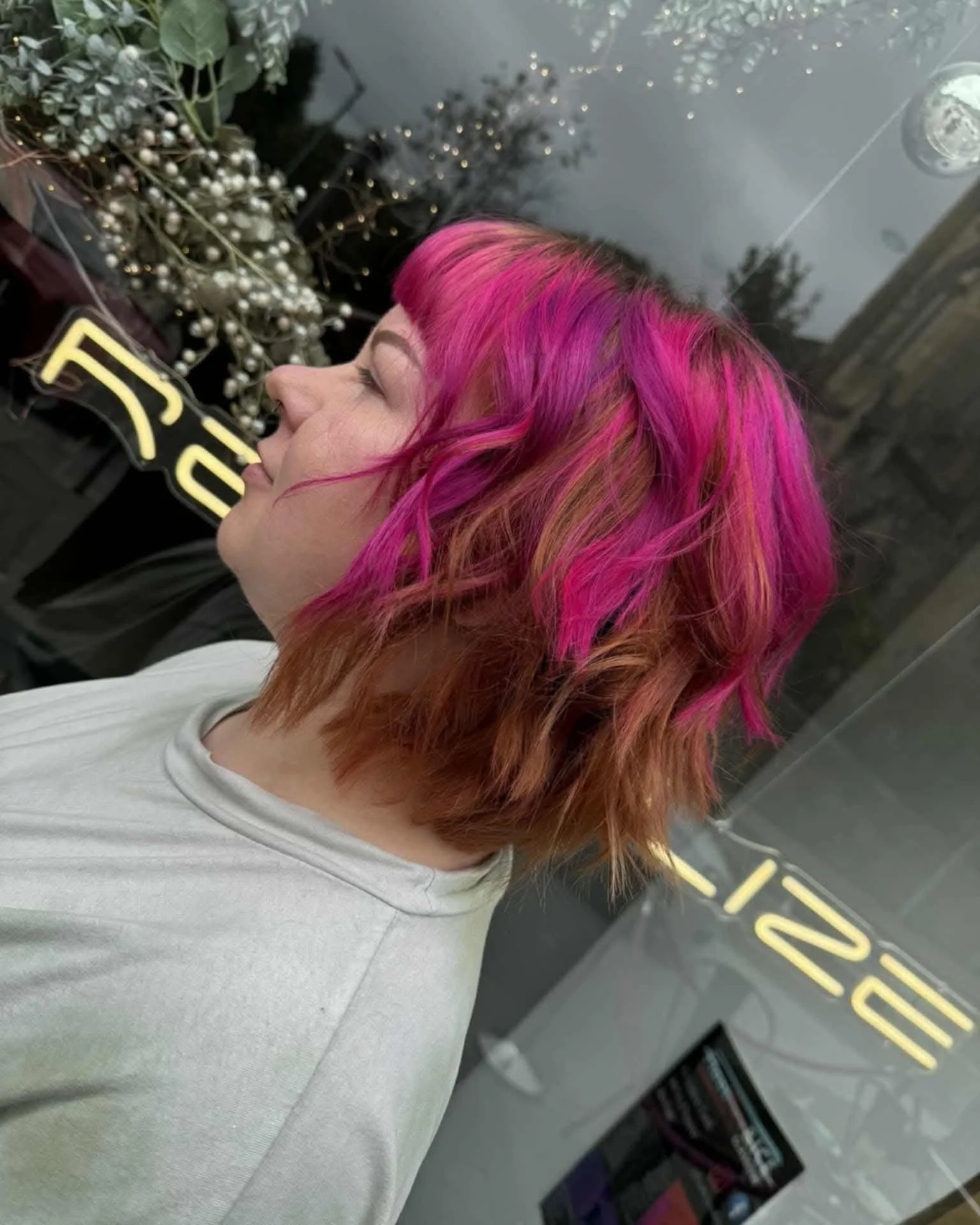 A woman with short hair dyed bright pink, standing in a salon or hair studio, with a background of a black wall with decorative pearly white berries and a neon sign.