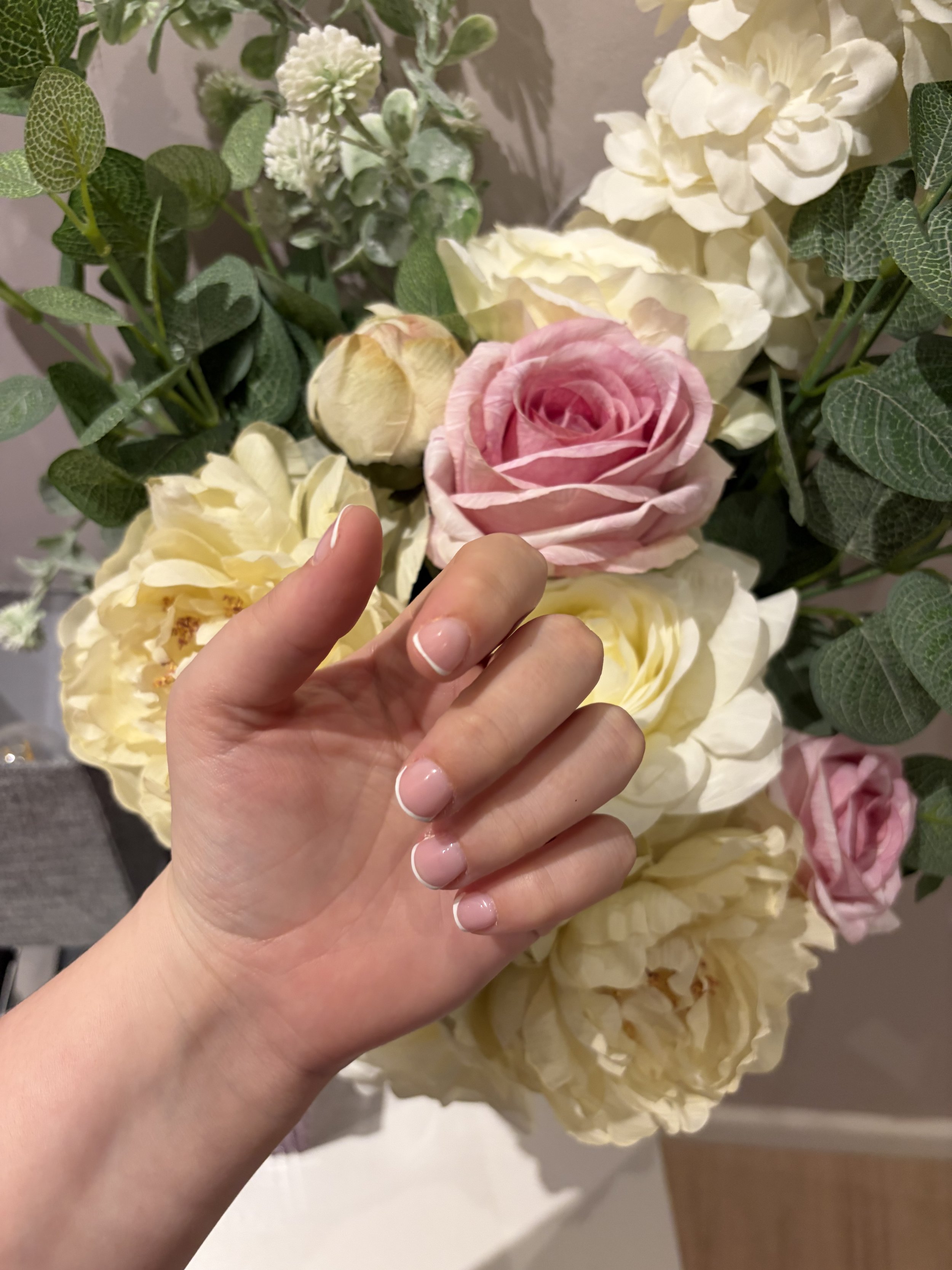 Close-up of a hand with well-manicured nails touching artificial flowers, including a pink rose, cream-colored roses, and other greenery.