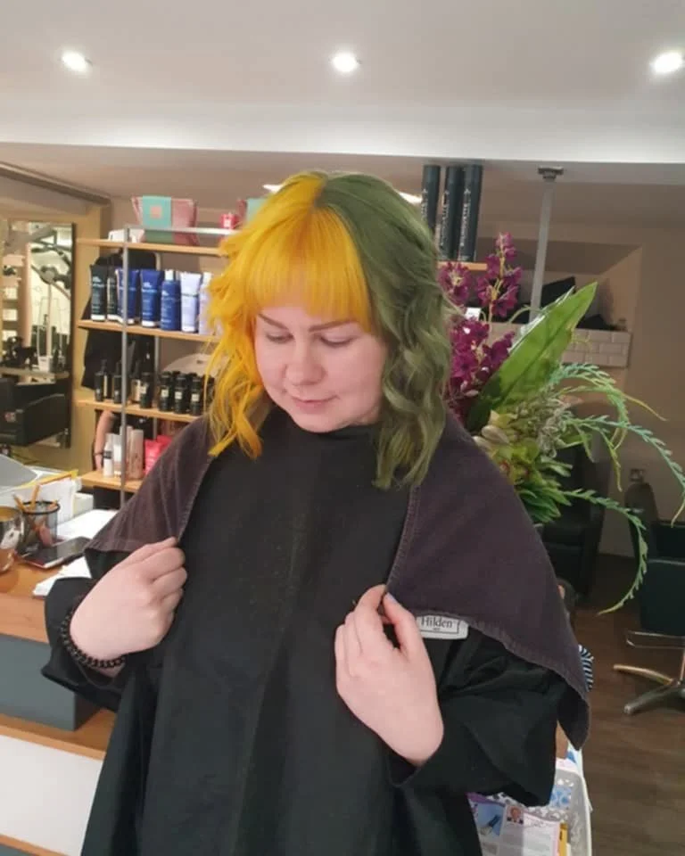 A person with half yellow and half green hair standing in a salon, wearing a black cape, with a background of shelves with hair products and a large purple flower arrangement.